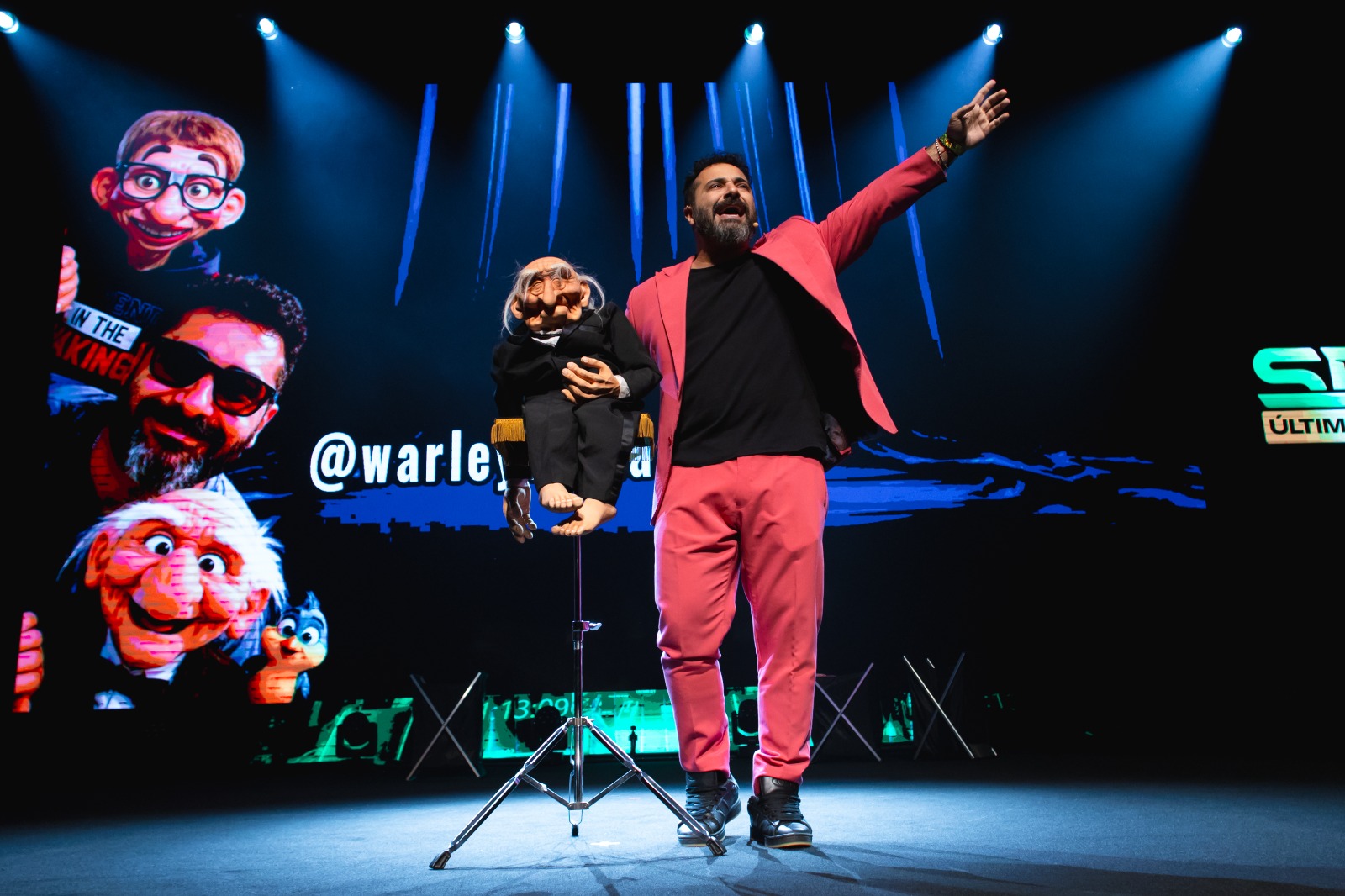 Man on stage with a puppet, raising his right arm, with a large screen behind displaying animated characters and text.