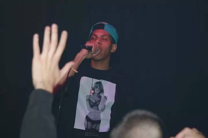 Person singing into microphone with raised hand in foreground, dark background, wearing a cap and graphic t-shirt