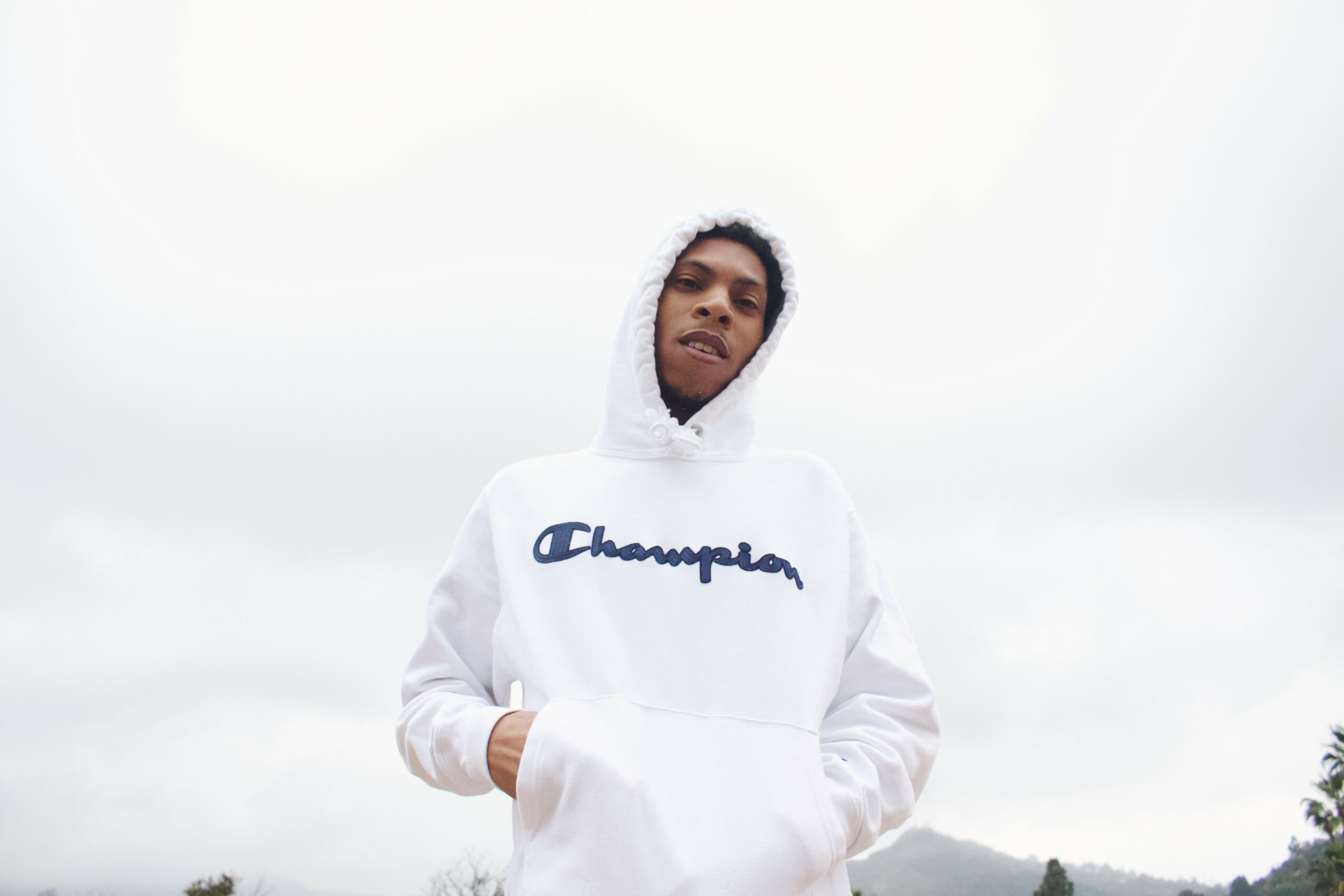 Young man wearing a white hoodie with a logo, standing outdoors against a cloudy sky.