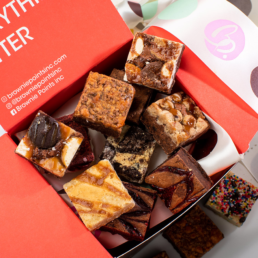 Assorted brownie squares with various toppings inside a red box, some with chocolate, nuts, and caramel, on white paper with colorful sprinkles.