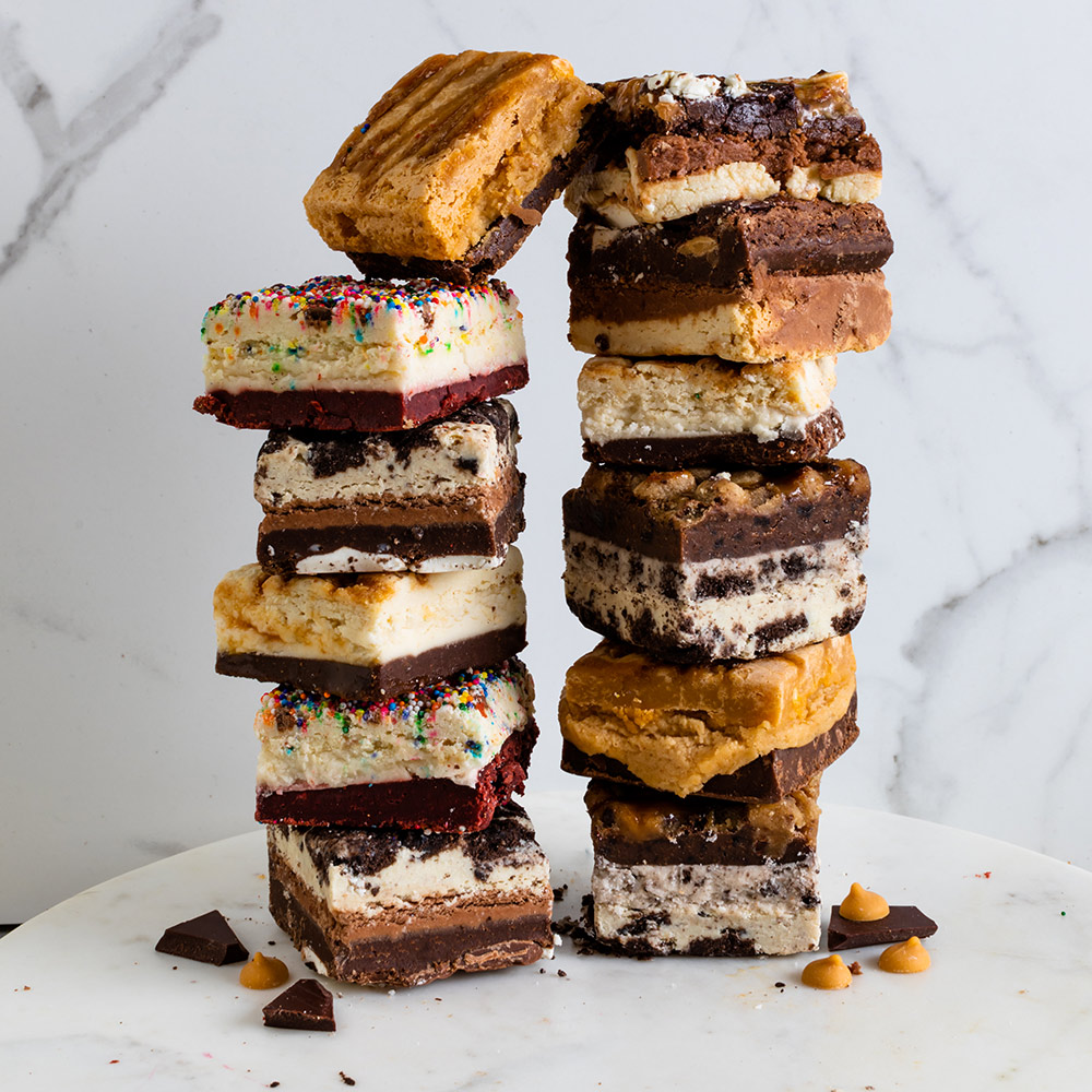 Two stacks of assorted brownies and cookies on a white surface, with a marble background, some chocolate pieces around.