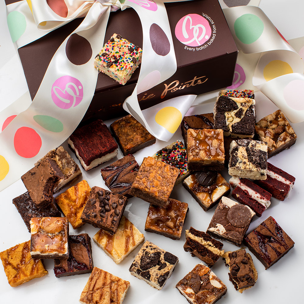 Assorted small brownies and cake squares scattered around a decorated box with a pink logo, colorful ribbons, and confetti.