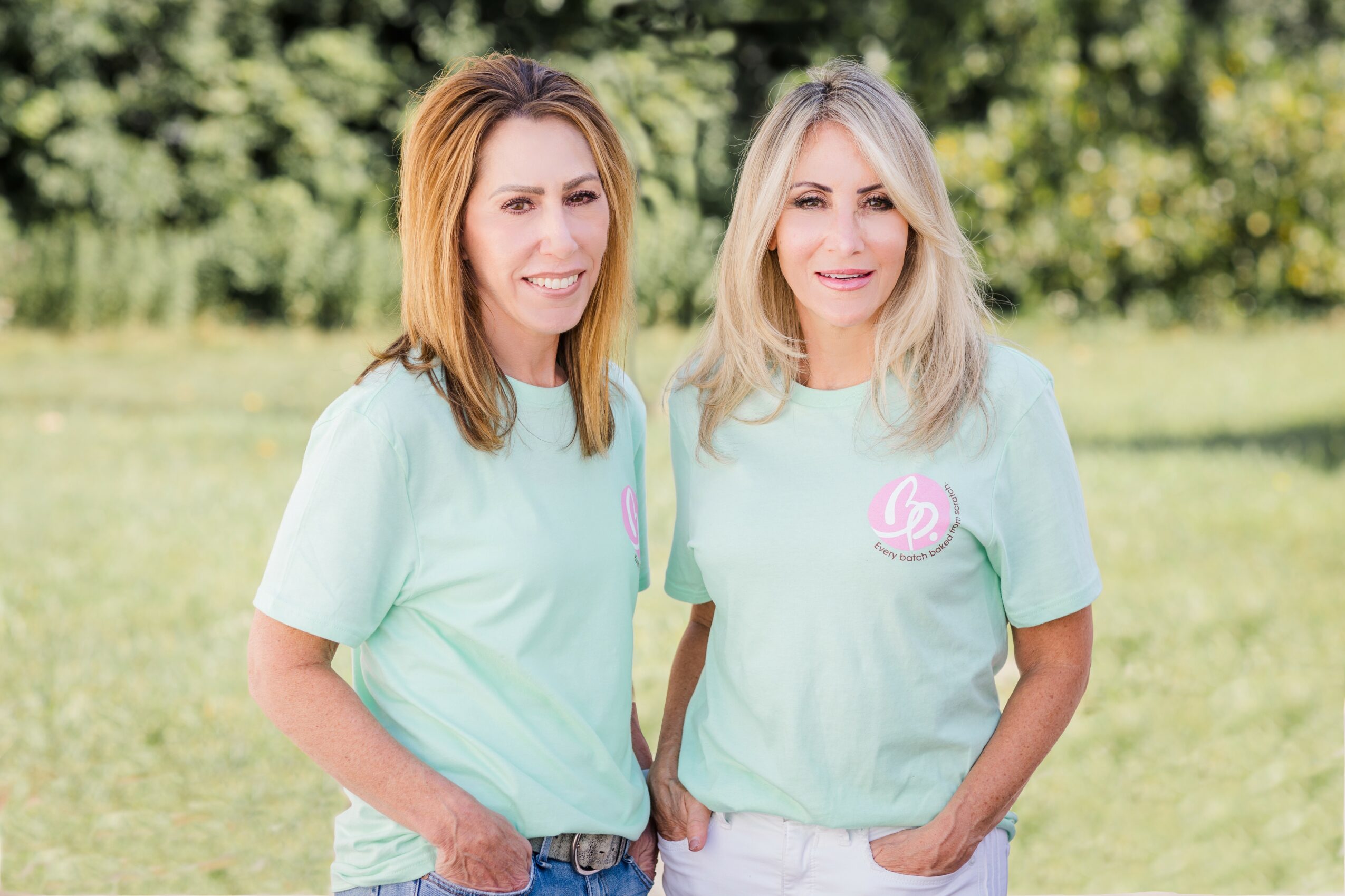 Two women standing outdoors on grass, smiling, wearing light-colored T-shirts, with trees in the background.