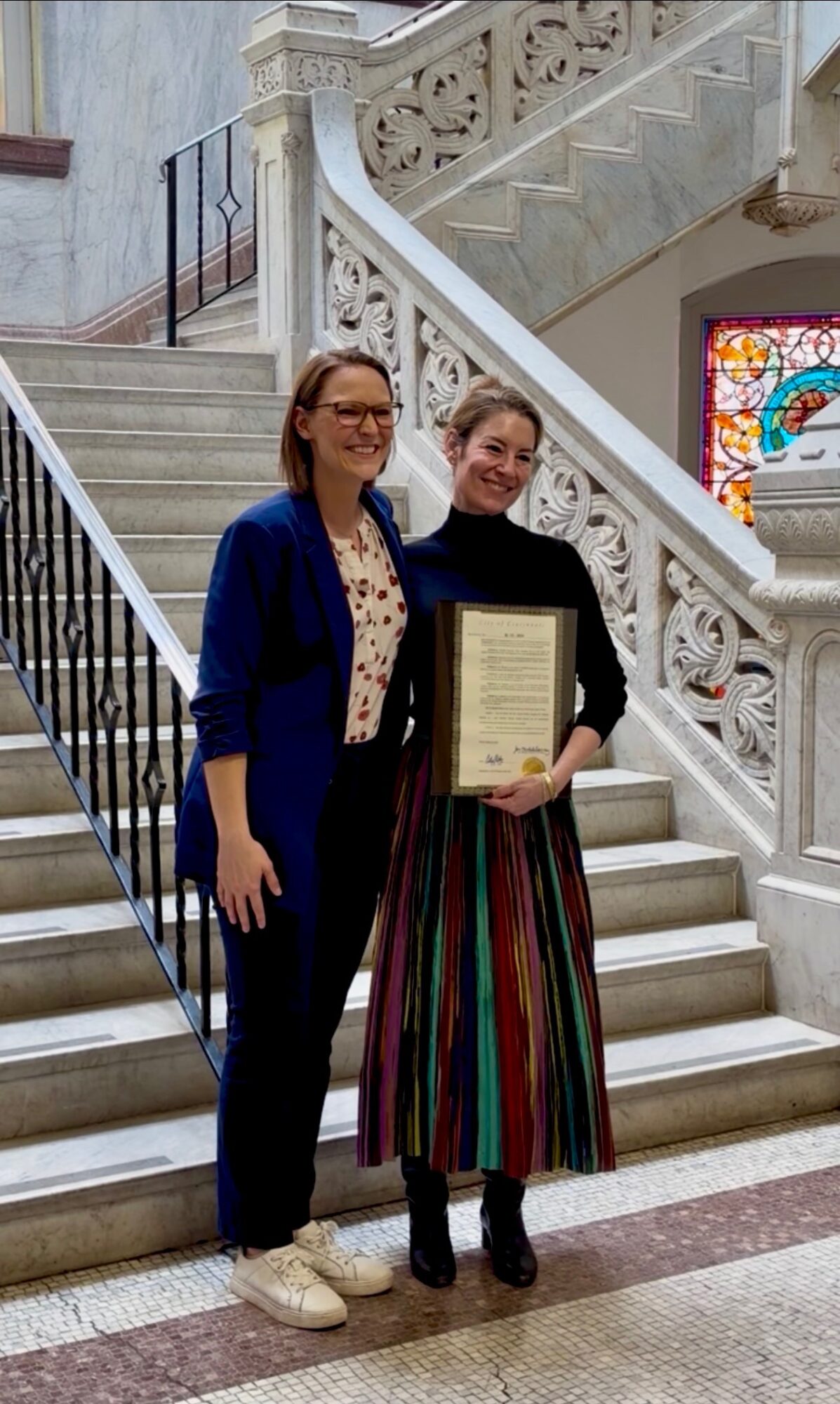 Two women standing on a staircase, one holding a framed certificate, in an elegant building with ornate railings and a stained glass window.