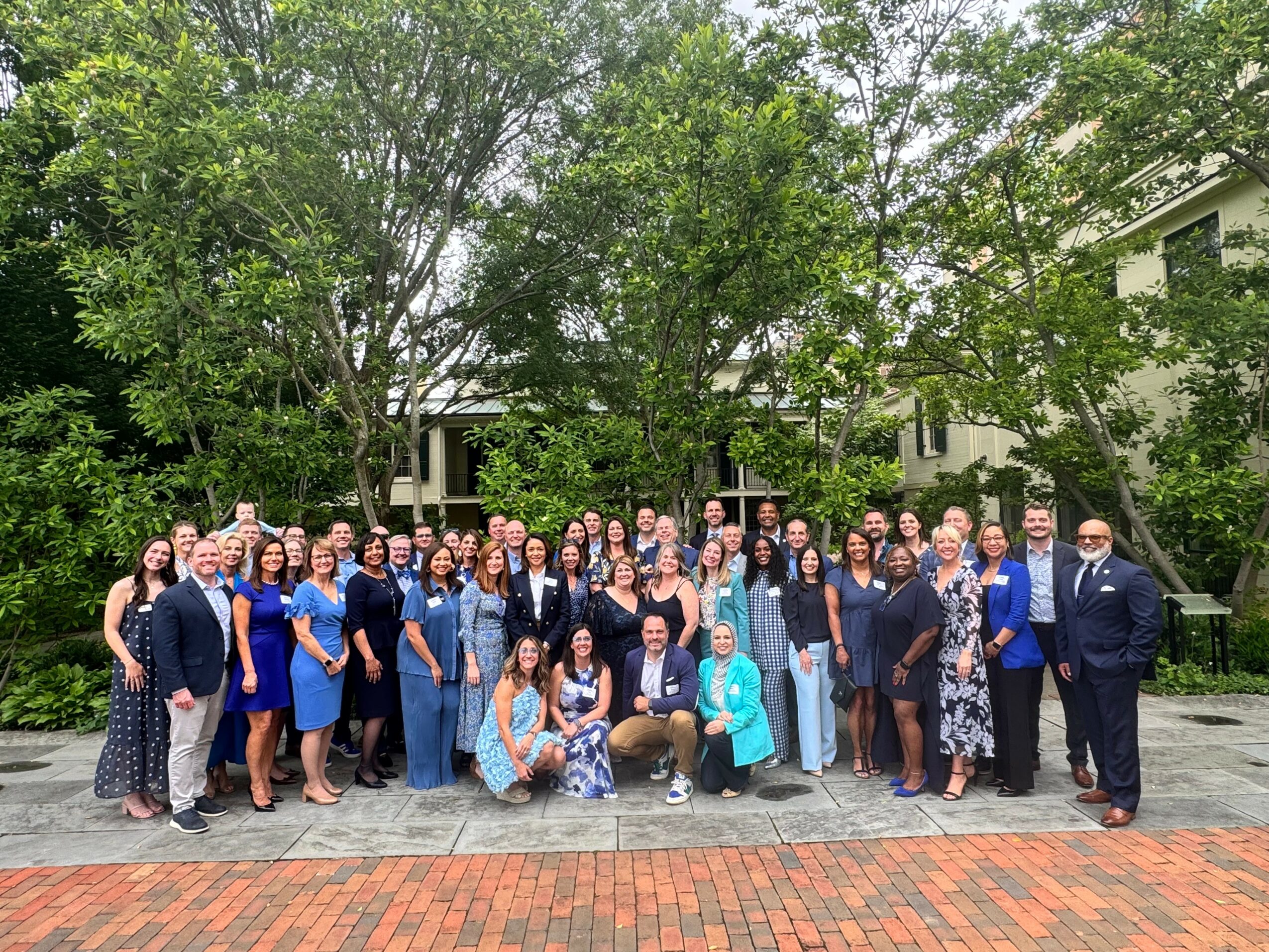 Group of people outdoors in front of trees and buildings, posing for a photo on a paved area, some smiling and standing close together.