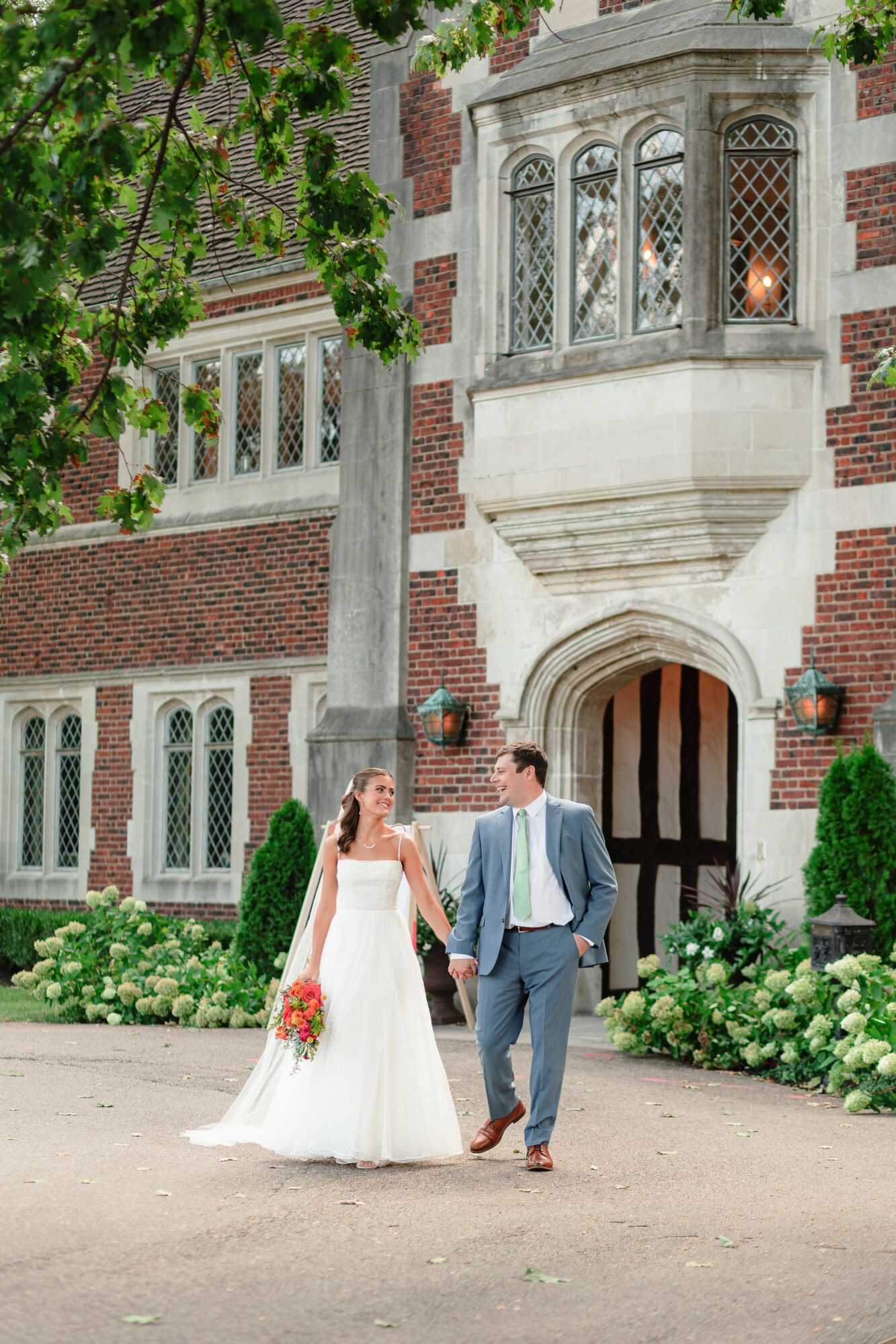 Bride and groom holding hands walking outside a historic brick building with large windows and decorative architecture, greenery around.