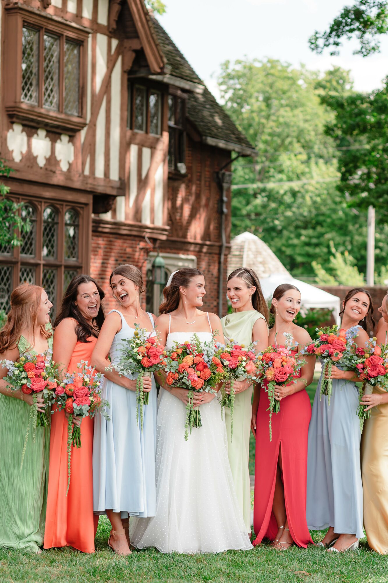 Group of women in colorful dresses holding bouquets outdoors in front of a historic building with trees in the background.