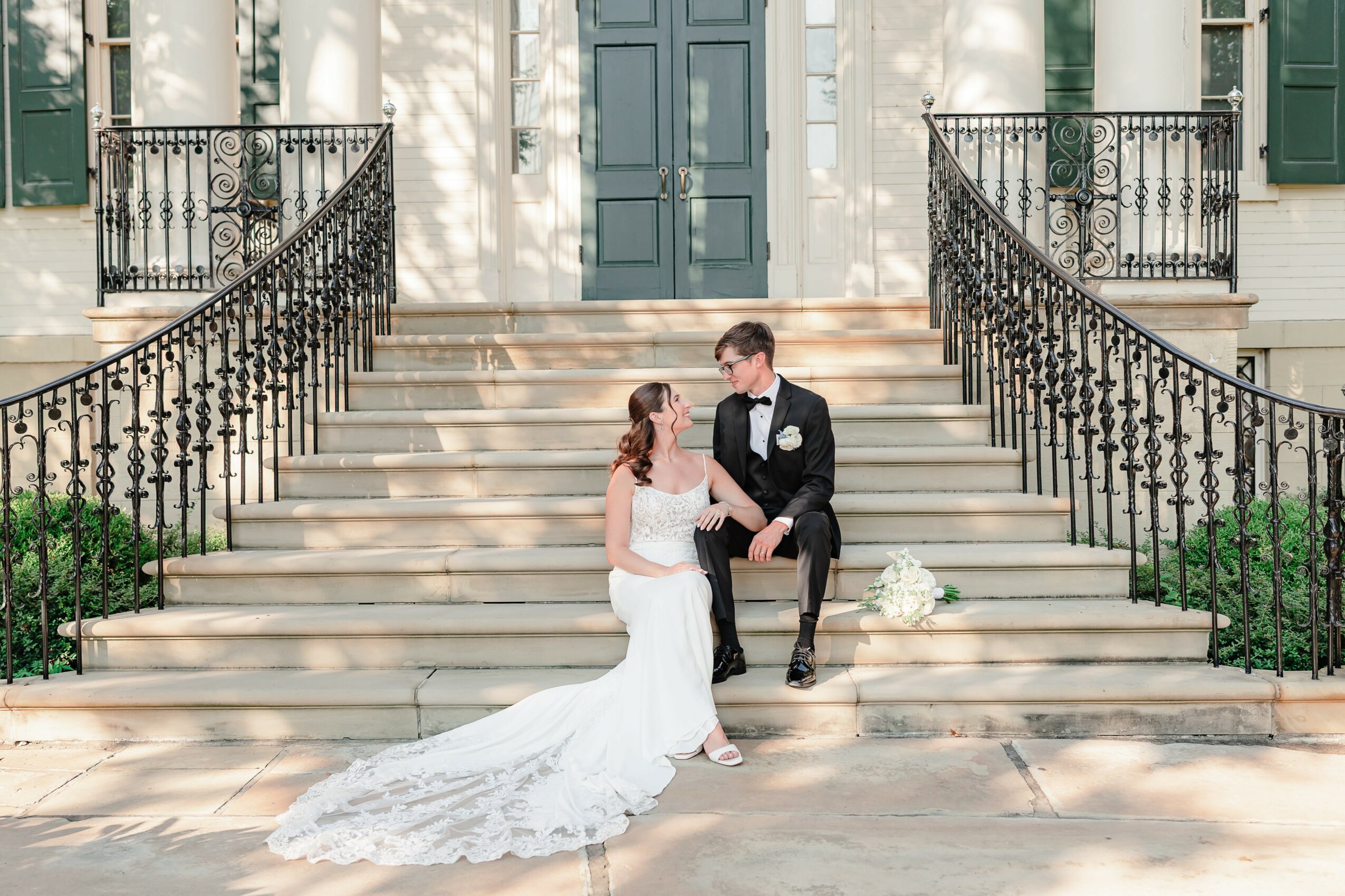 Bride and groom sitting on steps outside a building, holding hands, with wedding dress and bouquet, surrounded by black railings.
