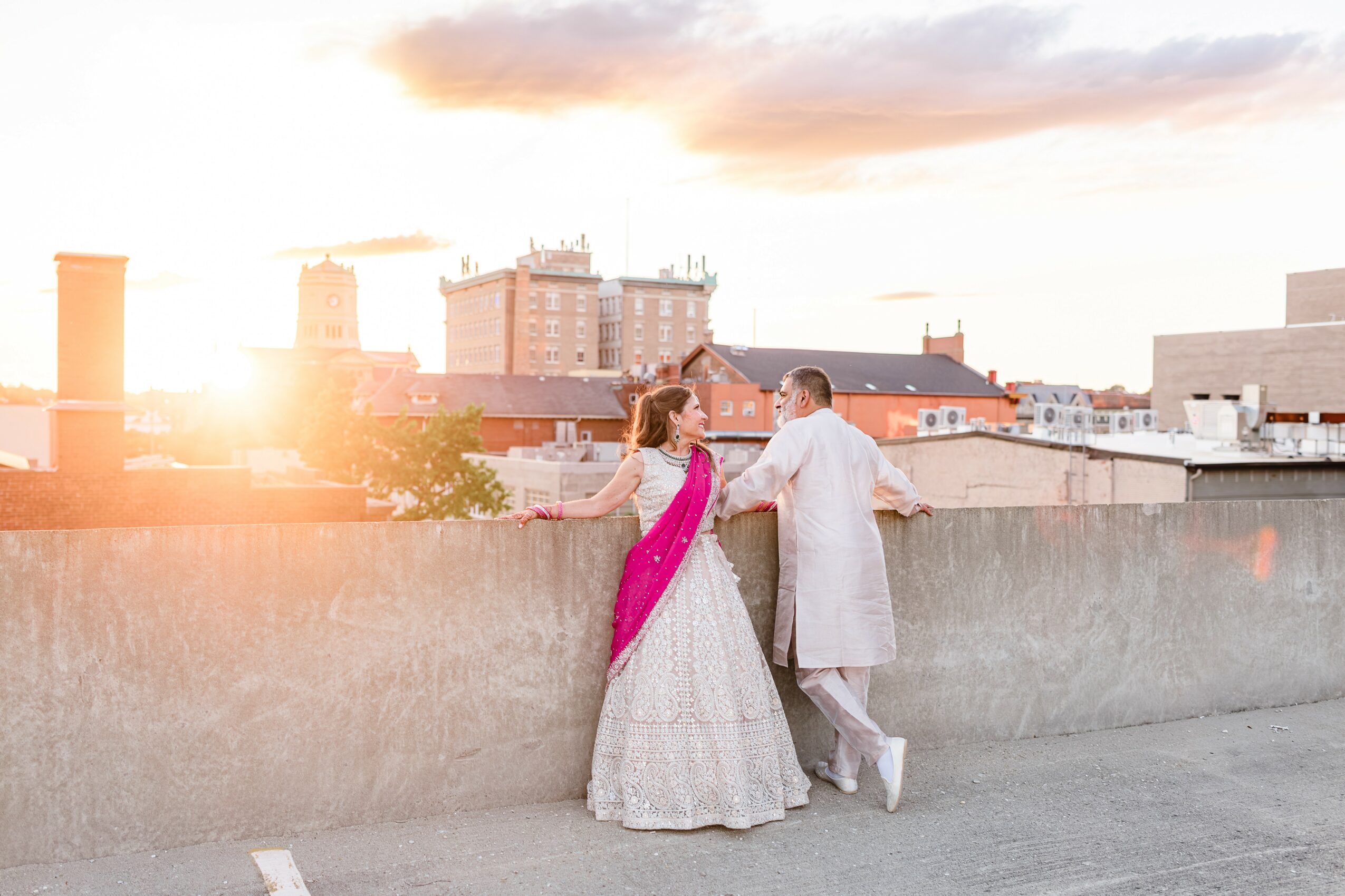 Two people in traditional attire stand on a rooftop at sunset, overlooking a cityscape with buildings and a cloudy sky.