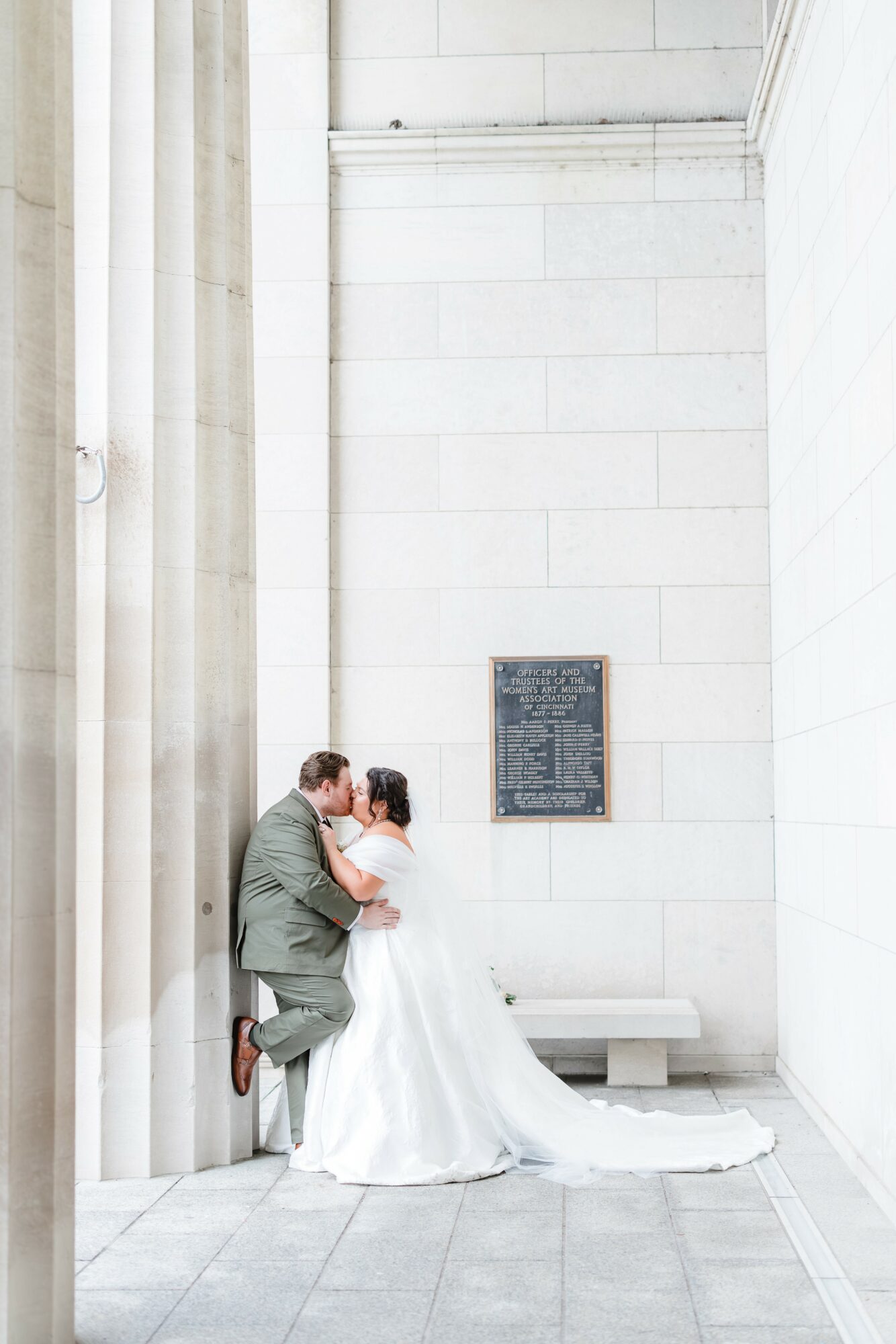 A couple in wedding attire kissing against a white wall with columns and a plaque, in a bright, minimal setting.