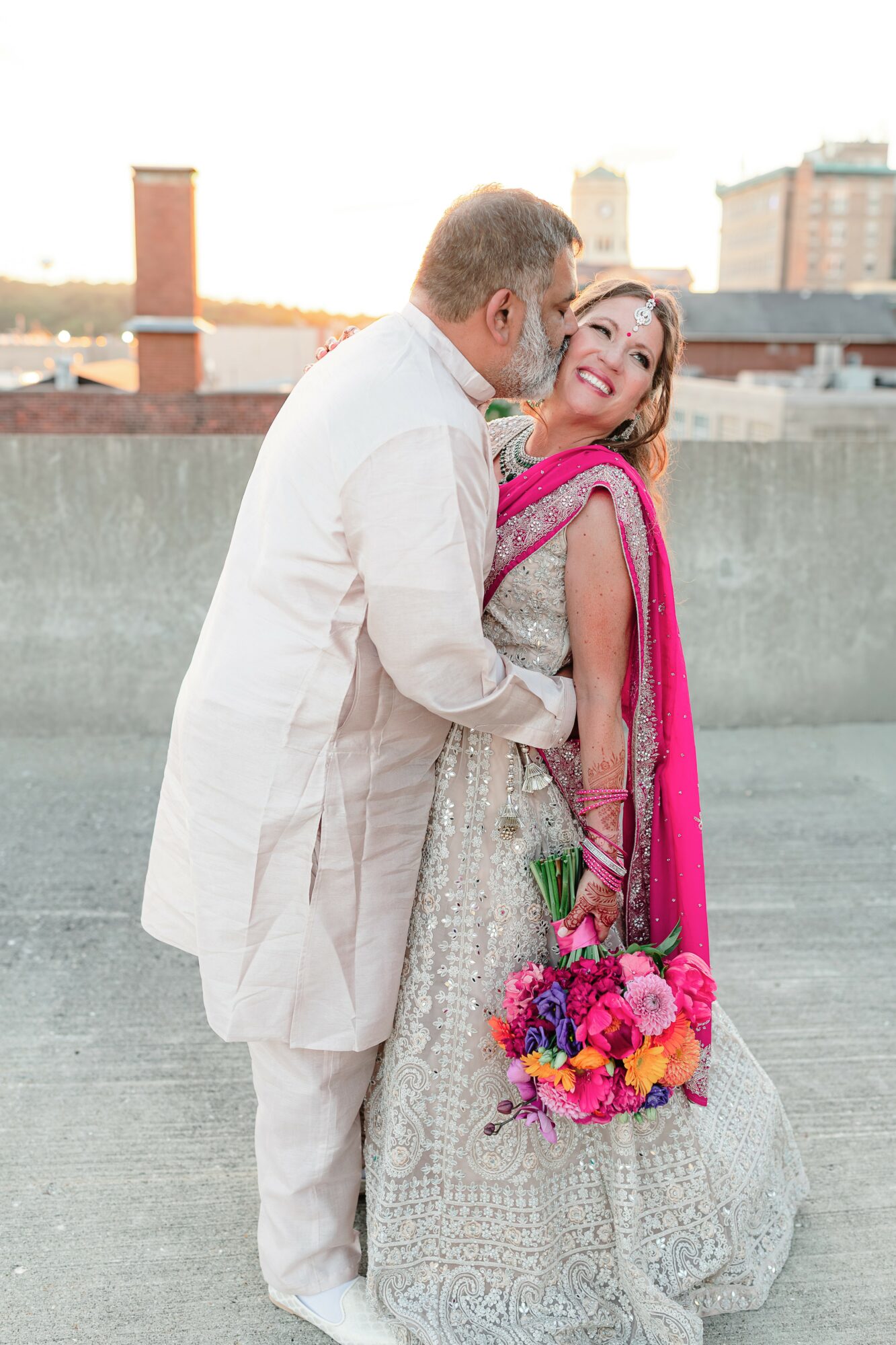A couple dressed in traditional attire on a rooftop, smiling and holding a colorful bouquet of flowers.