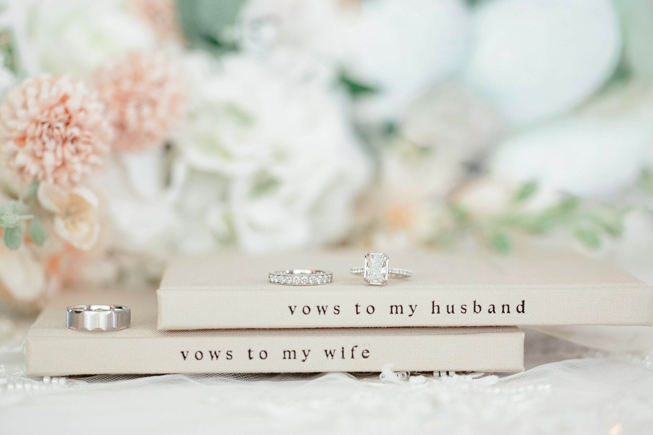 Two wedding vow books, one labeled for the husband and the other for the wife, with rings on top, in front of flowers