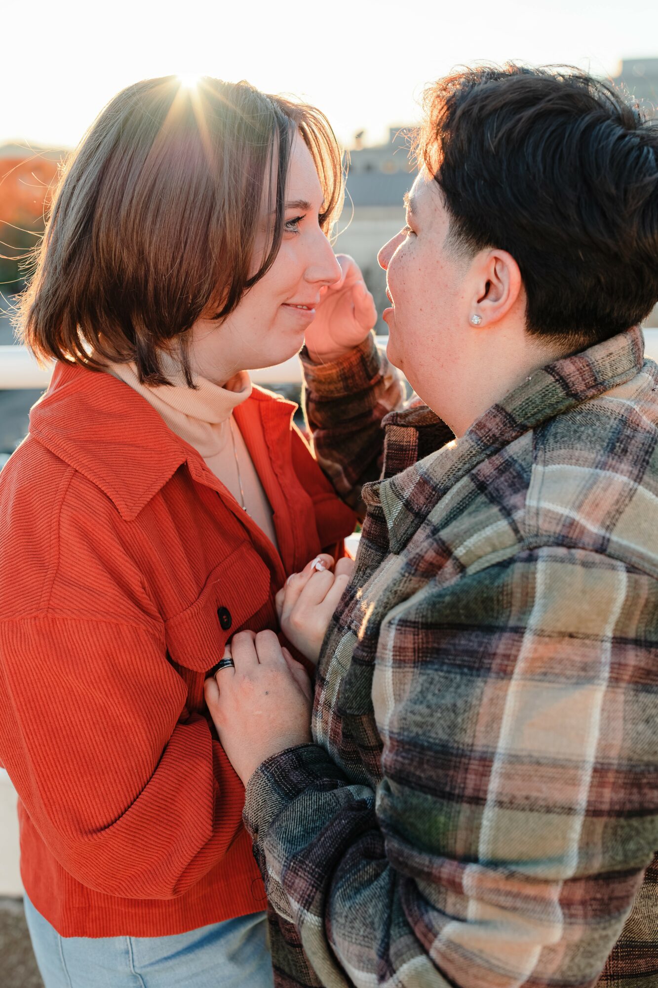 Two people face each other outdoors, close together, with sunlight behind them, holding hands and smiling.