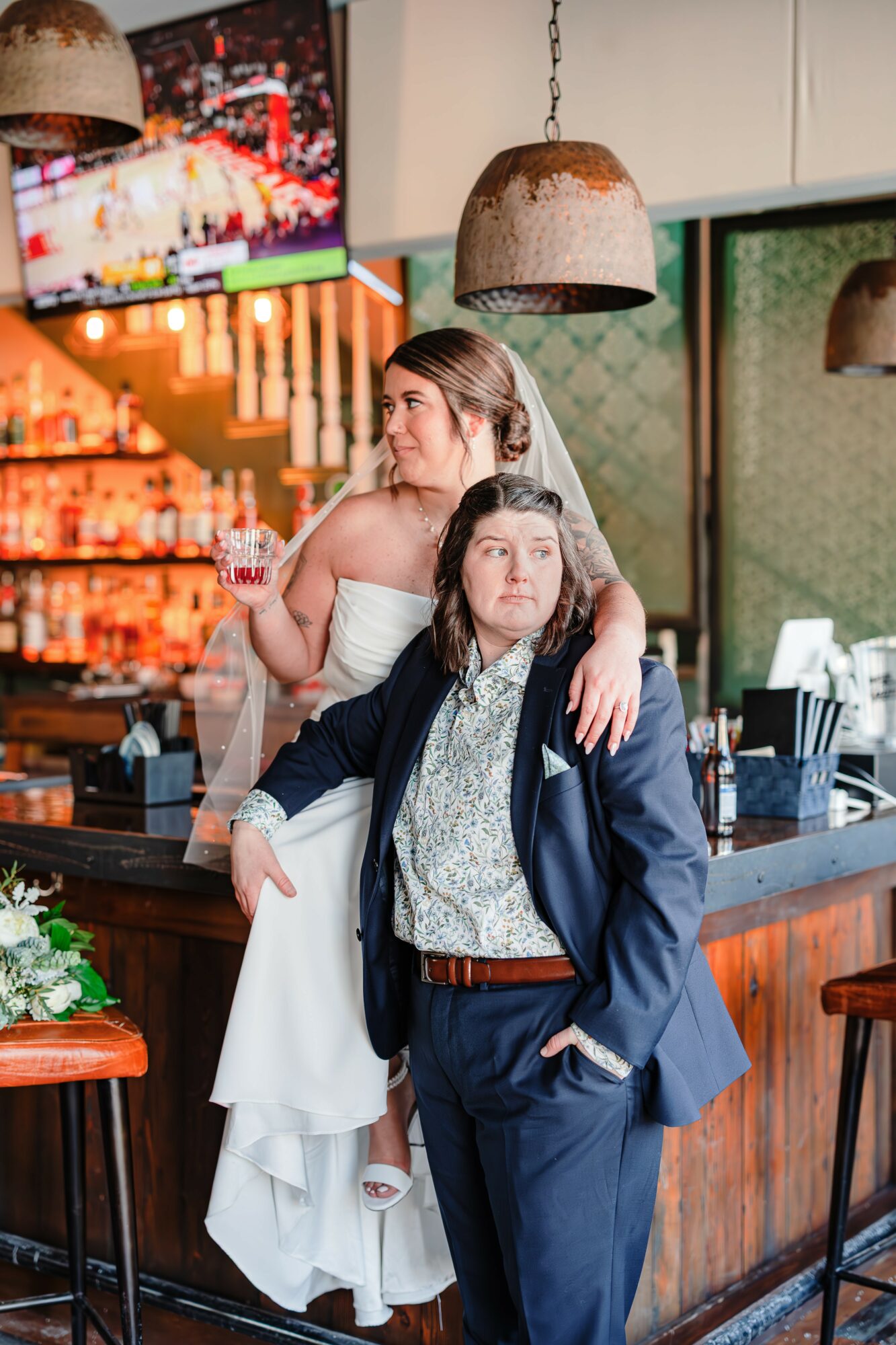 Two women, one in a wedding dress and the other in a suit, stand at a bar with a TV and hanging lights behind them.