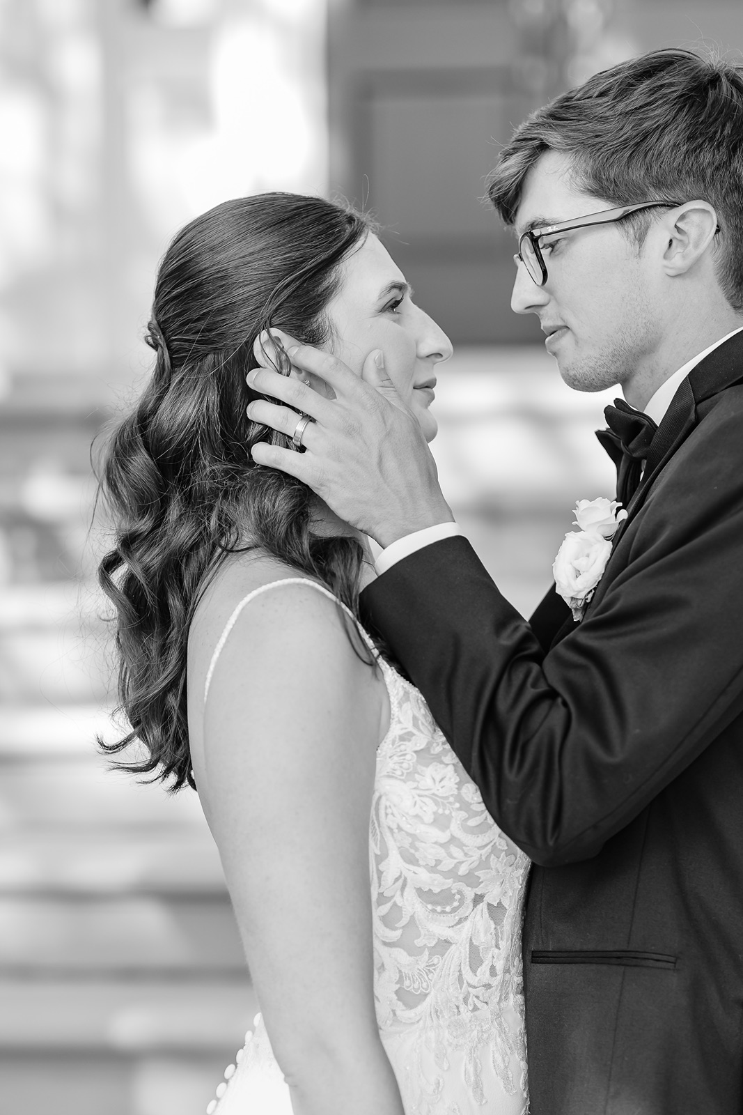 A couple in wedding attire gazes at each other, with the man gently holding the woman's face, in black and white.
