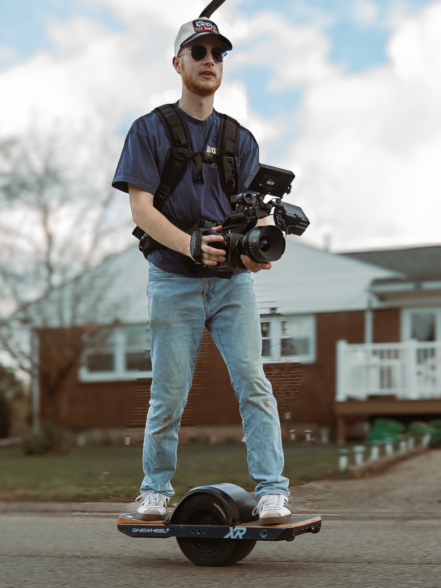 Young man standing on a hoverboard outdoors, holding a camera, wearing sunglasses, a cap, and casual clothes.