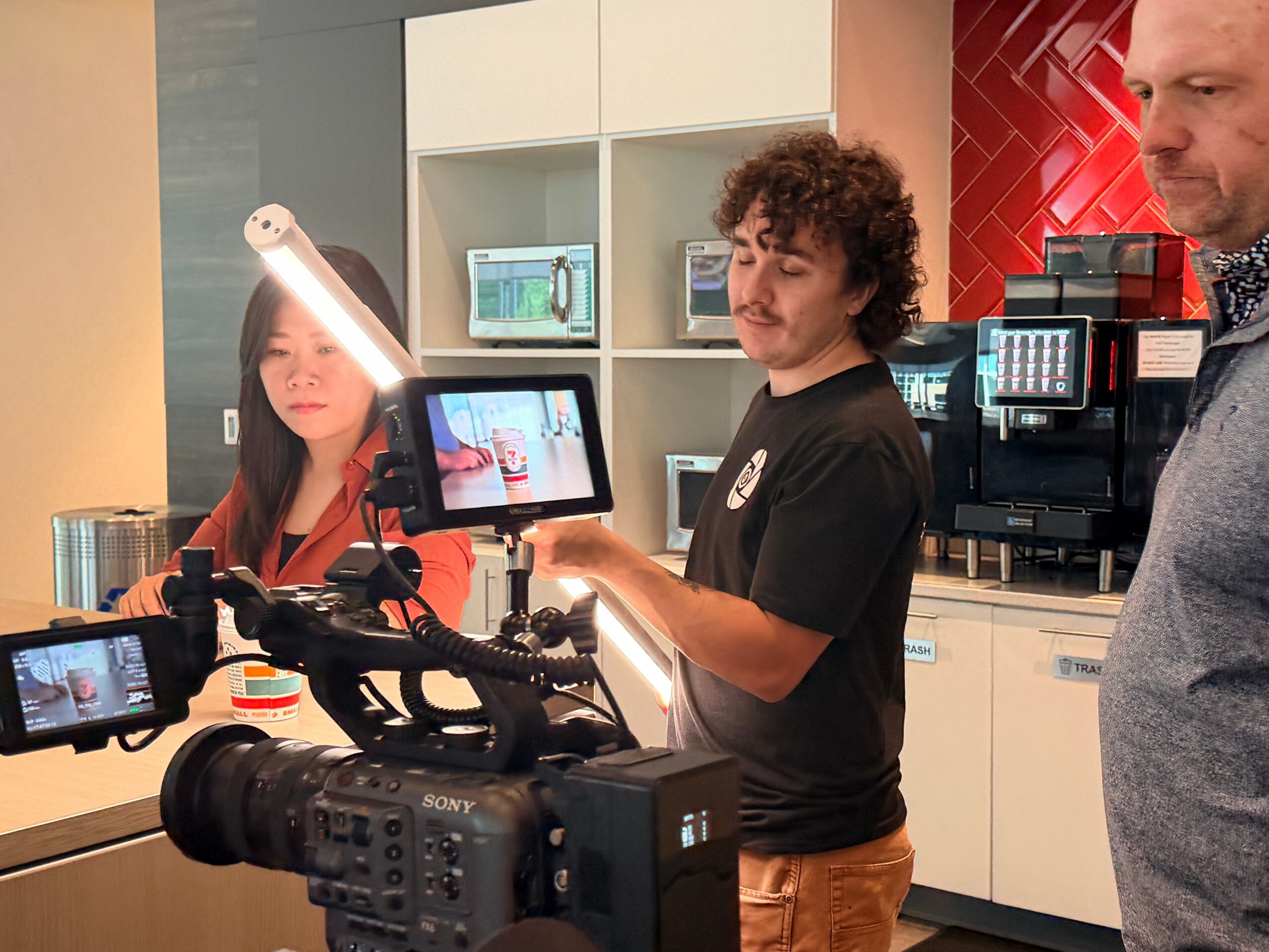 Three people in a kitchen, one person operating a camera, two others observing, with kitchen appliances in the background.