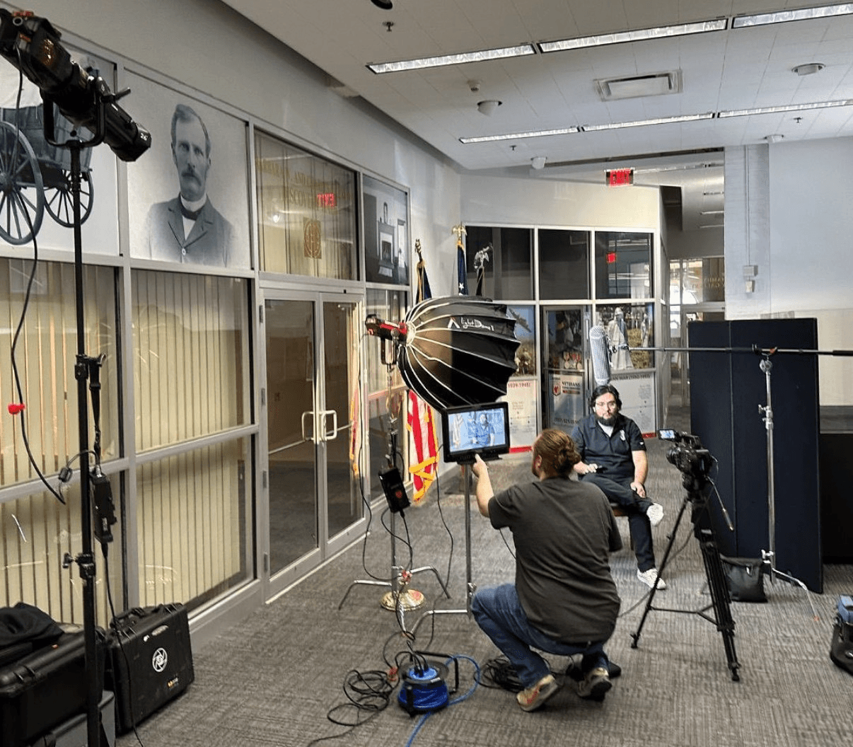 Person being interviewed in a room with cameras and lighting equipment, portraits on the wall, and a person filming.
