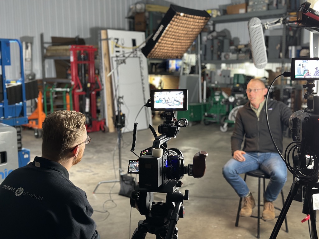 Person being interviewed sits on a stool in a workshop with equipment and machinery in the background.