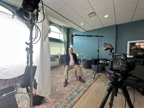 Man sitting on a chair in a room with blue walls, surrounded by professional camera and lighting equipment, during a video shoot.