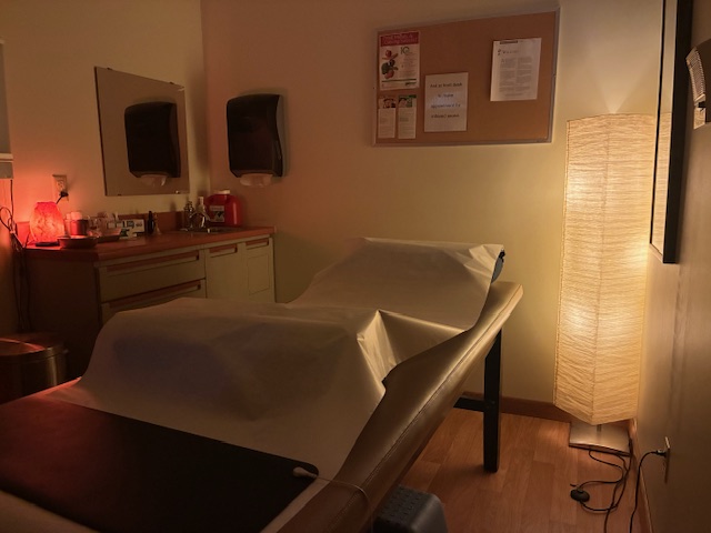 Hospital or clinic examination room with a bed, bulletin board, and a tall lamp, illuminated by warm lighting.
