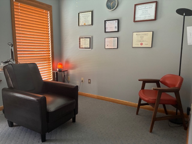 Office corner with armchair, wooden window blinds, framed certificates, and a red chair, with a floor lamp and small table.