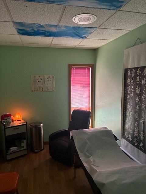 Room with green walls, window with pink blinds, table with tablecloth, and a small lamp on a cabinet.
