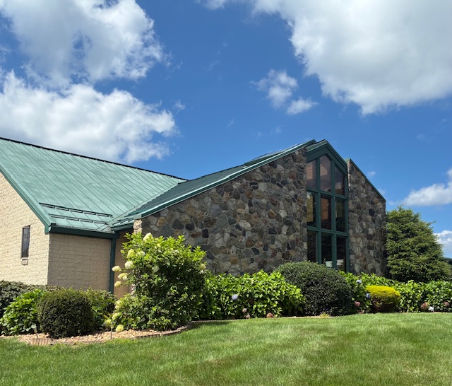 Building with stone and brick walls, large windows, green roof, surrounded by bushes and grass, under a partly cloudy sky.