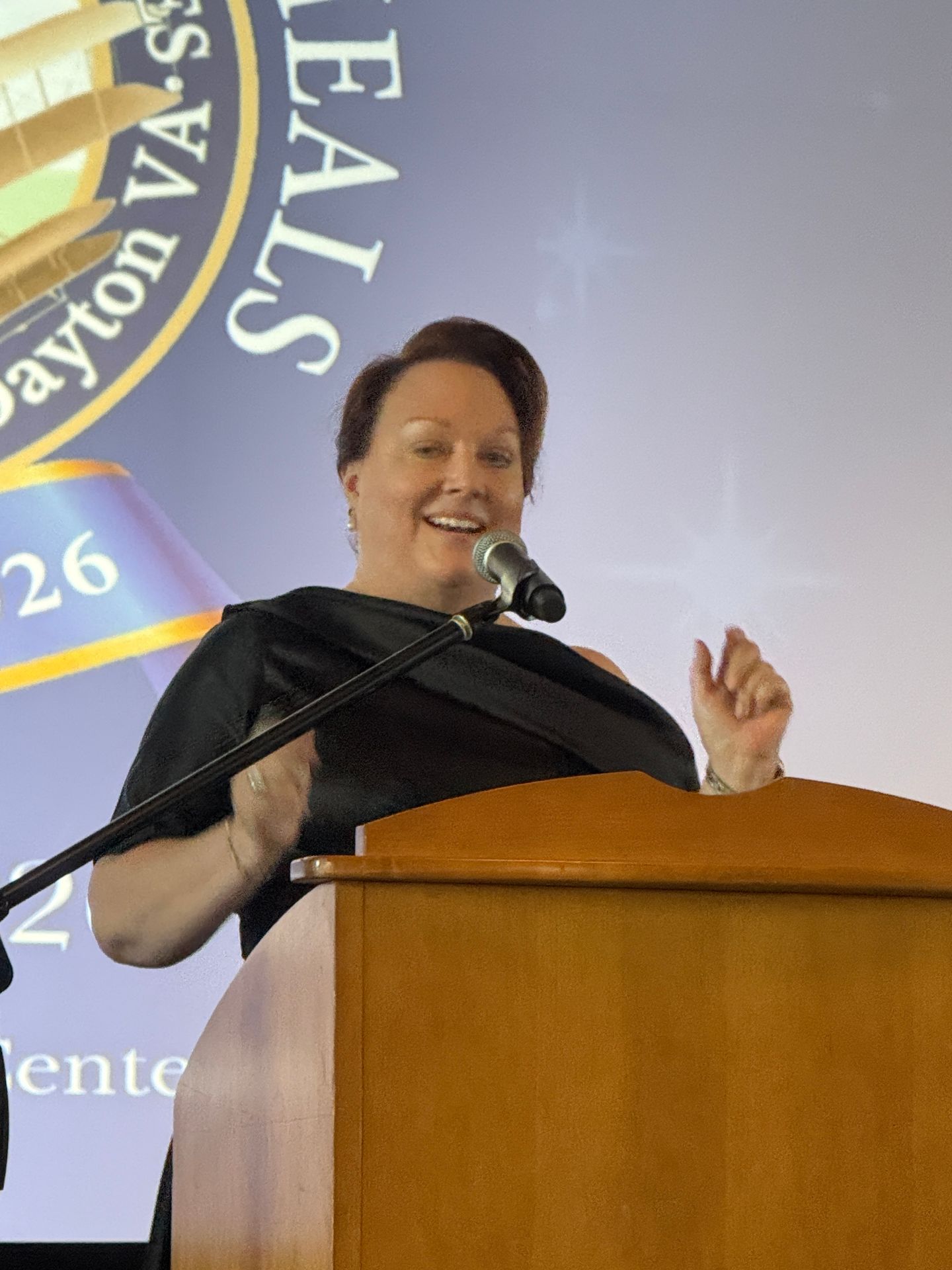 Woman speaking at a podium with a microphone, smiling, with a blue background and a logo behind her.