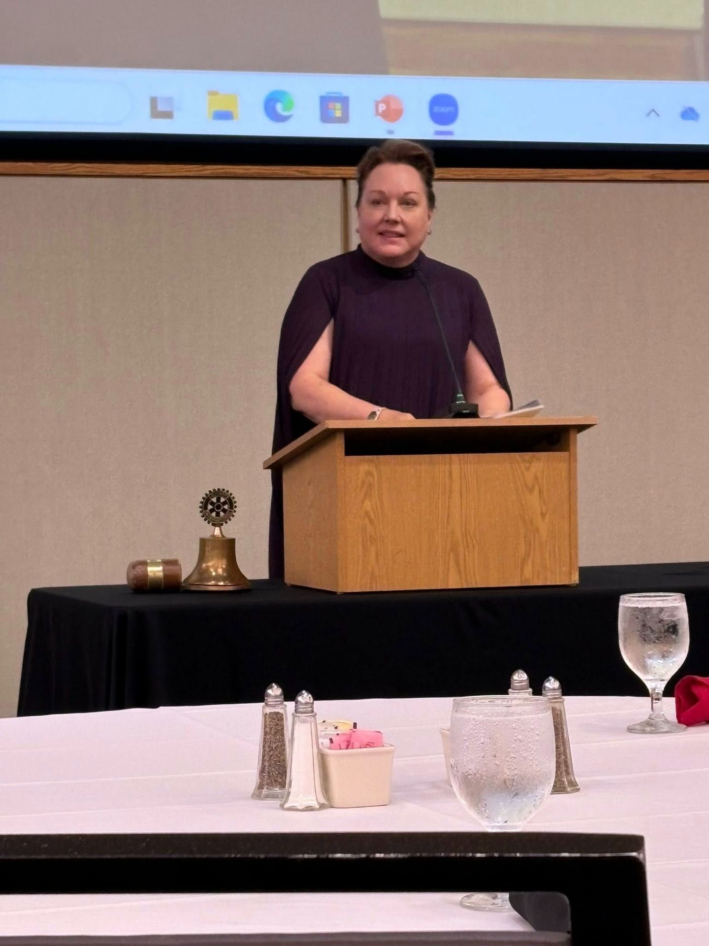 Woman standing behind a wooden podium, speaking in a conference room with a large screen behind her, table with drinks and items in foreground.