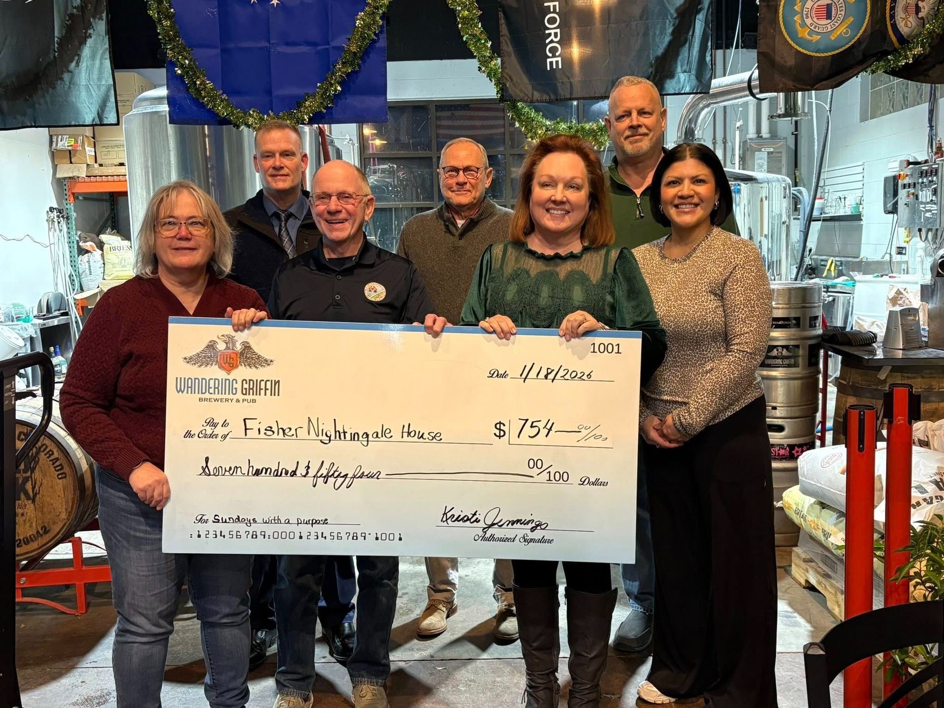 Group of seven people holding a large check inside a restaurant, smiling, with decorations and kitchen in background.