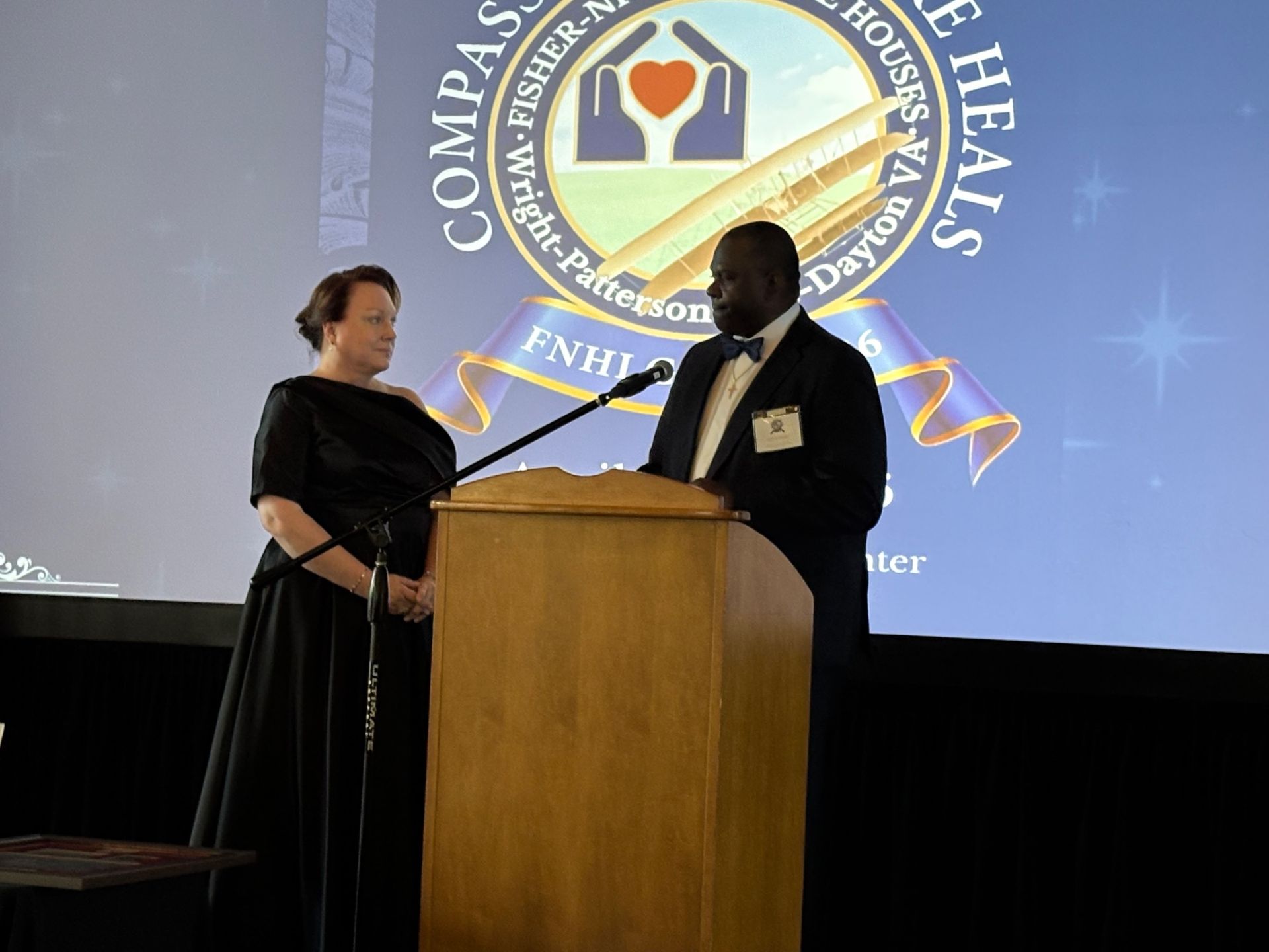 Two people standing at a podium on stage, with a large screen behind them displaying a logo and text.