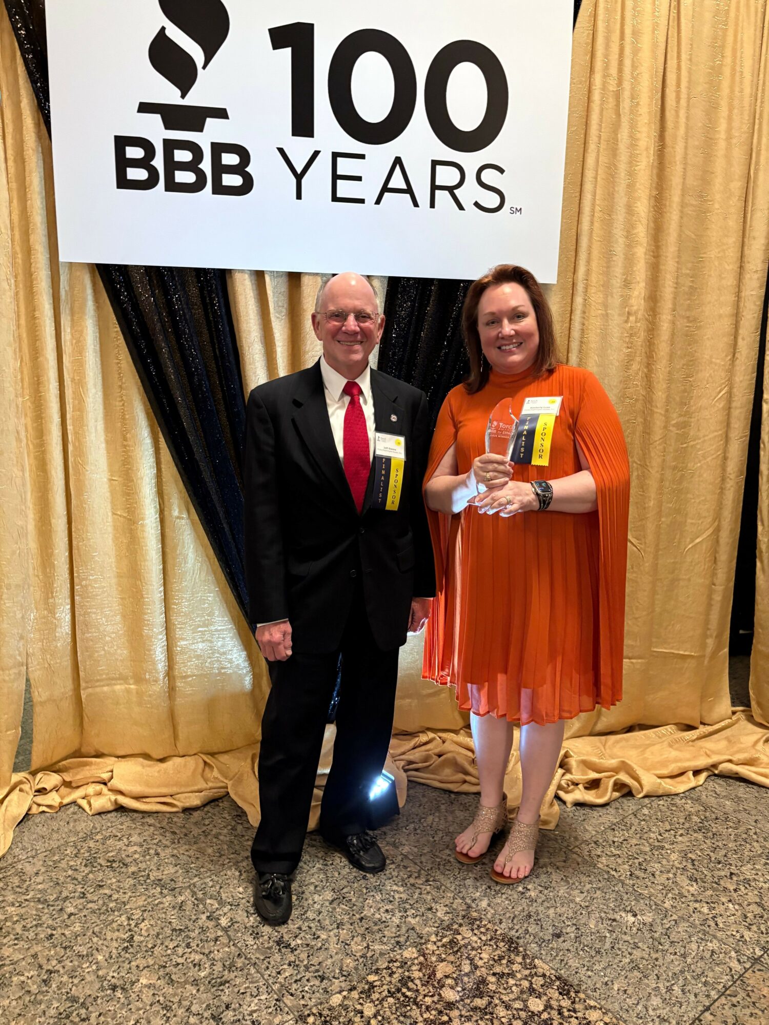 Two people standing in front of a beige curtain, holding a large sign that reads '100 BBB YEARS.' The person on the left wears an orange dress, and the person on the right wears a black suit with a red tie.