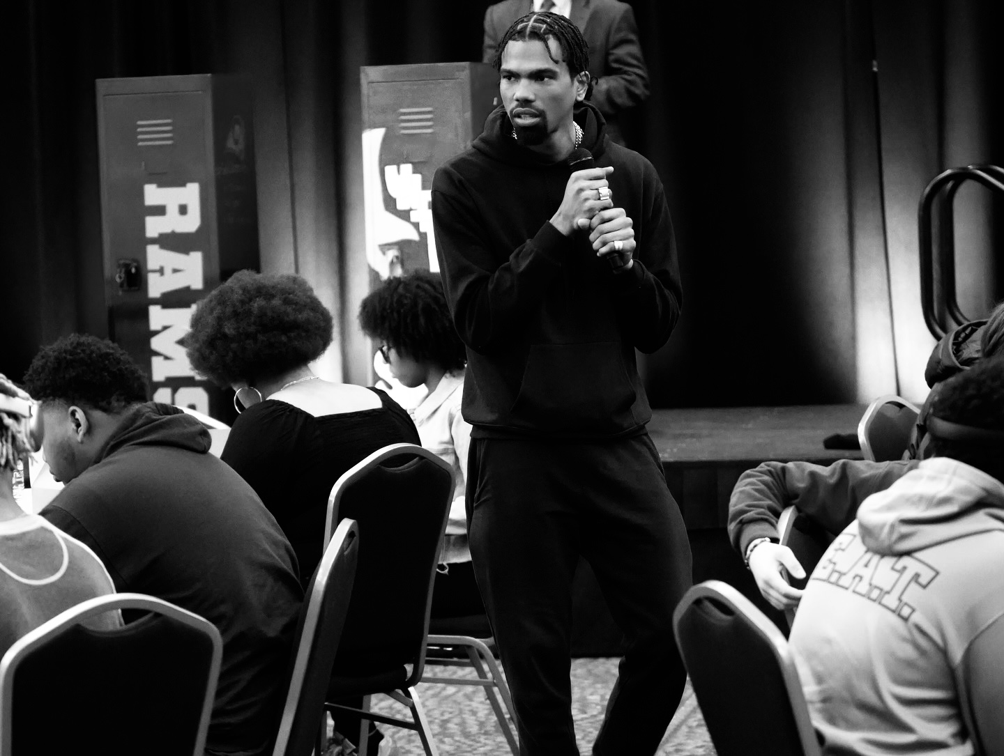 Man standing and speaking to an audience in a room with seated people, stage, and RAM banners, black and white photo.