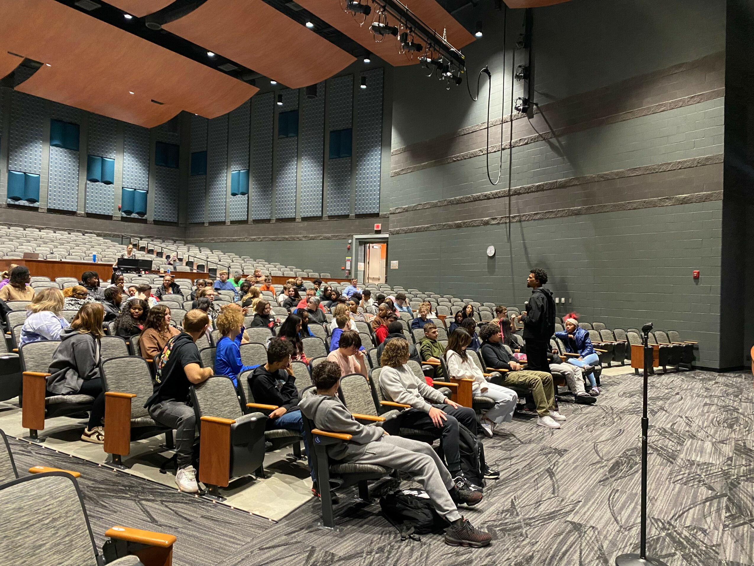 Audience seated in a large auditorium listening to a speaker on stage, with a high ceiling and wall panels.