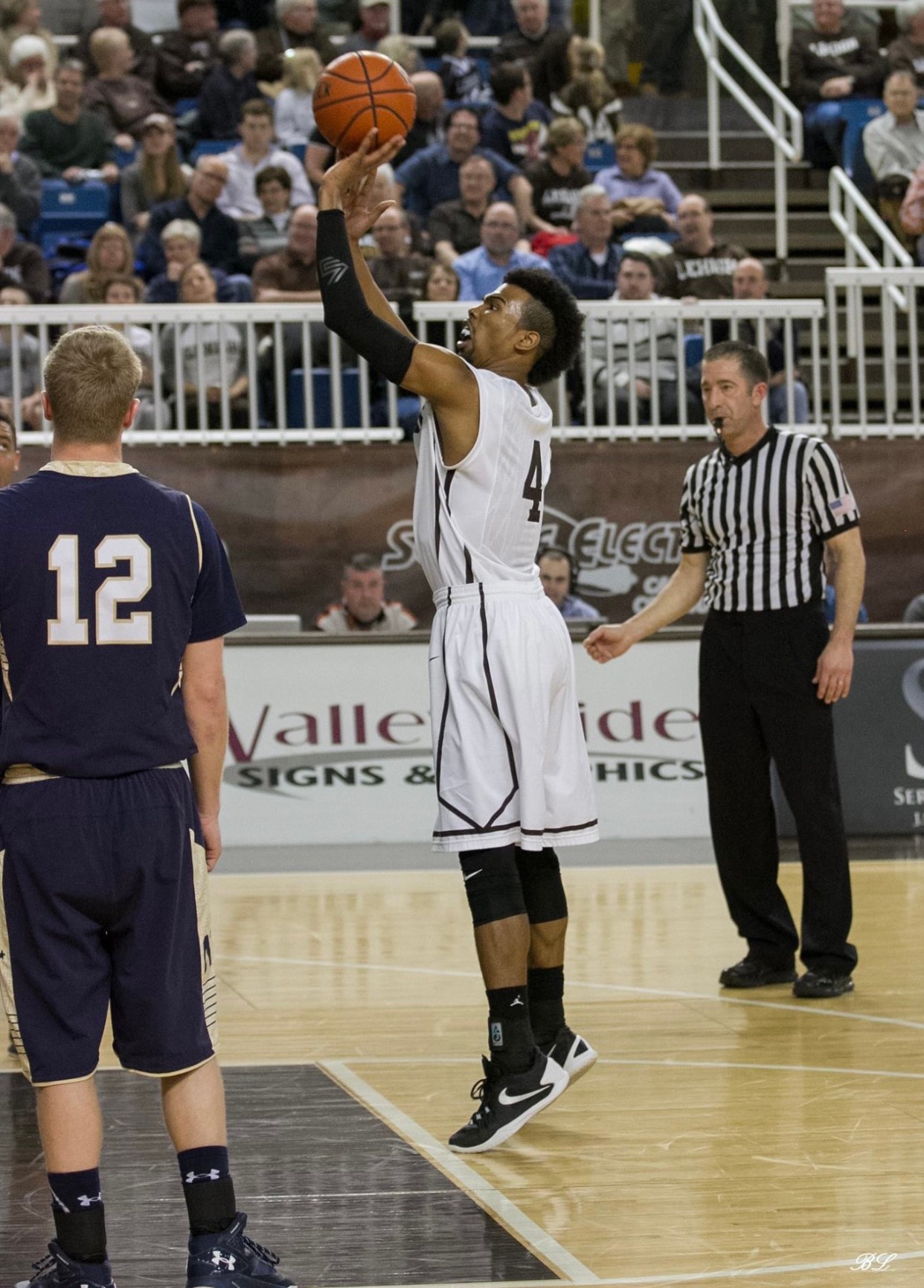 Basketball player in white uniform jumps to shoot, with referee and opponent nearby, audience in background.
