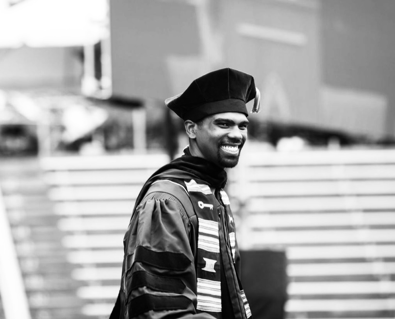 Smiling man wearing academic regalia and a cap, standing outdoors with blurred background.