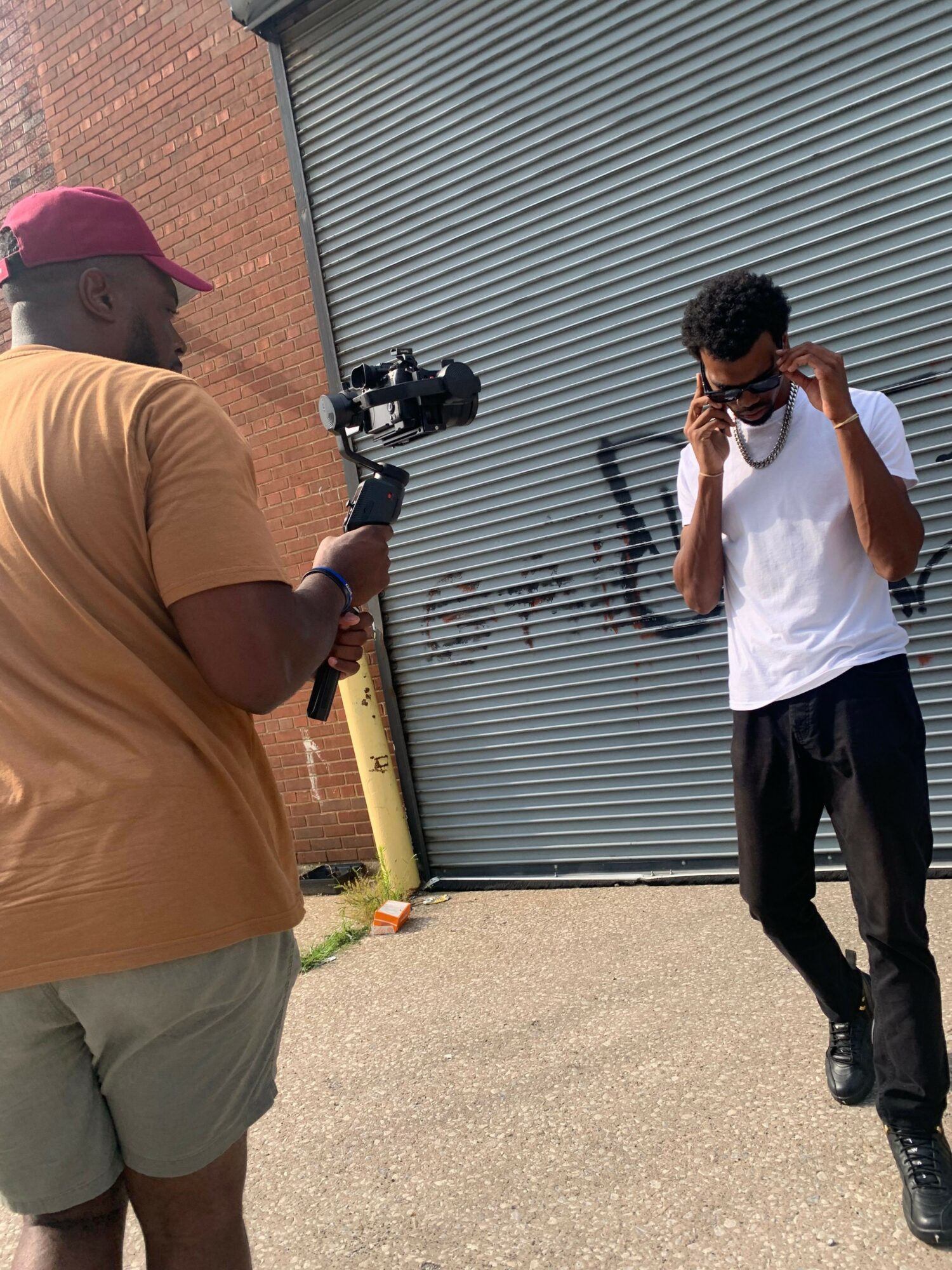 Person in white shirt and black pants walking past a gray metal shutter door, carrying a bag, with another person in brown shirt and shorts nearby.