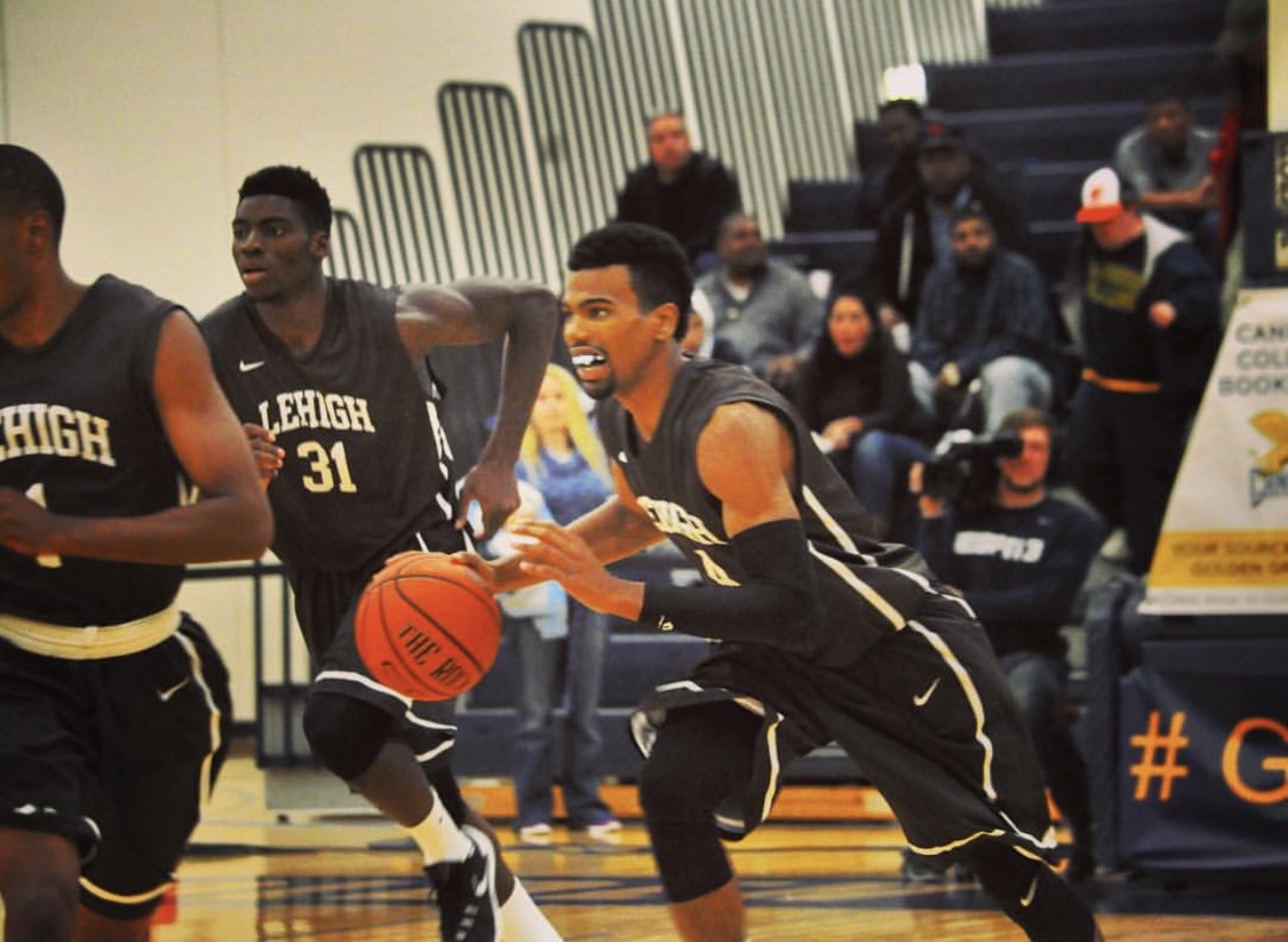 Basketball player dribbling during a game, with spectators in the background, some wearing masks, in an indoor gymnasium.