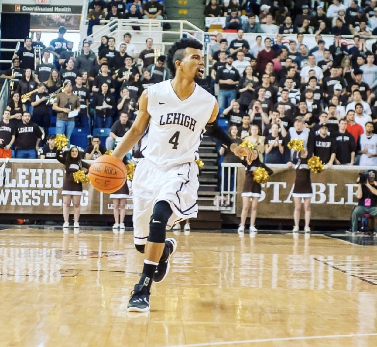 Basketball player in a white uniform dribbling on the court with a crowd and cheerleaders in the background.
