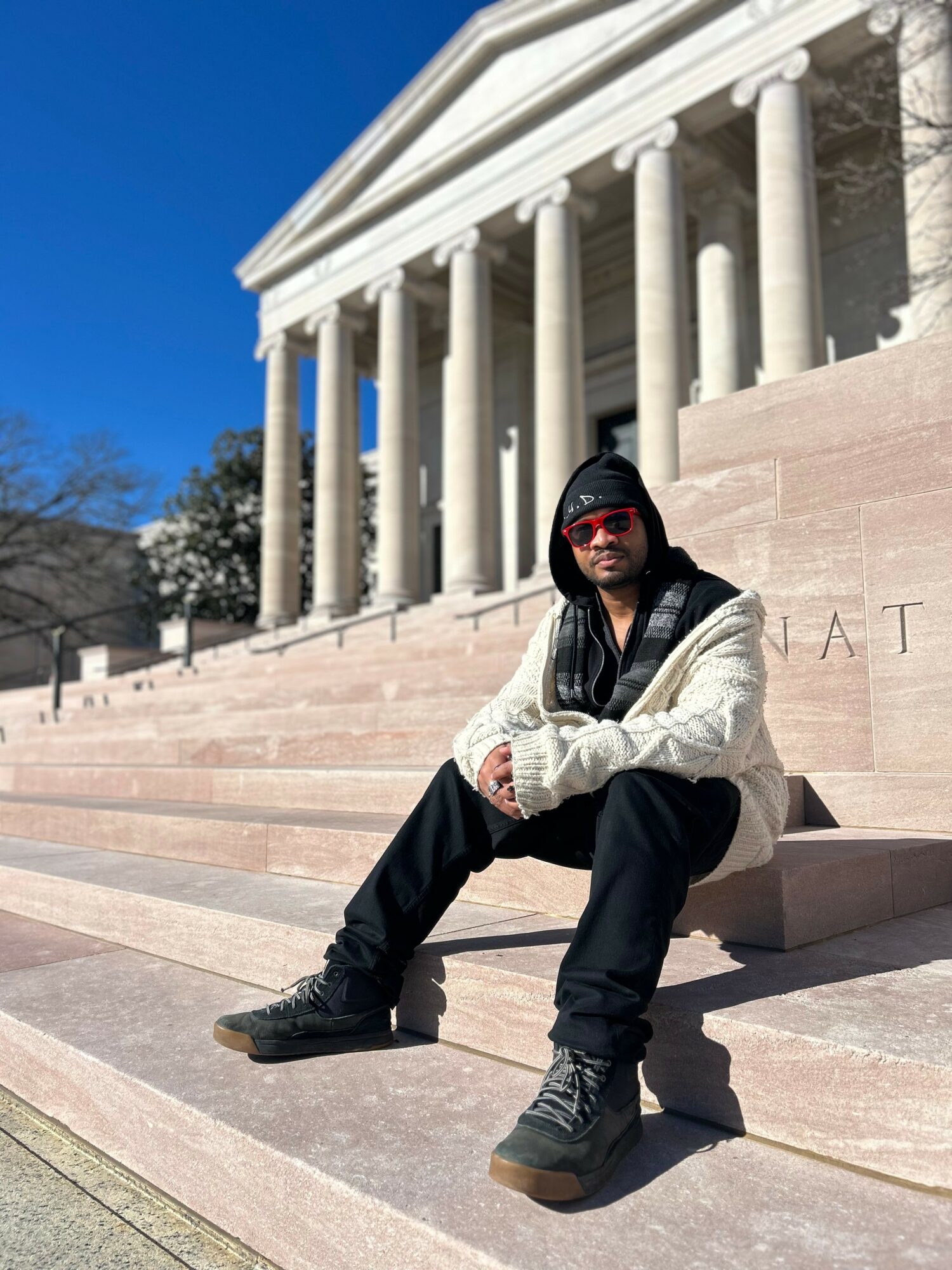 Person sitting on steps in front of a large classical building with columns, wearing a hoodie, sunglasses, and casual clothing.