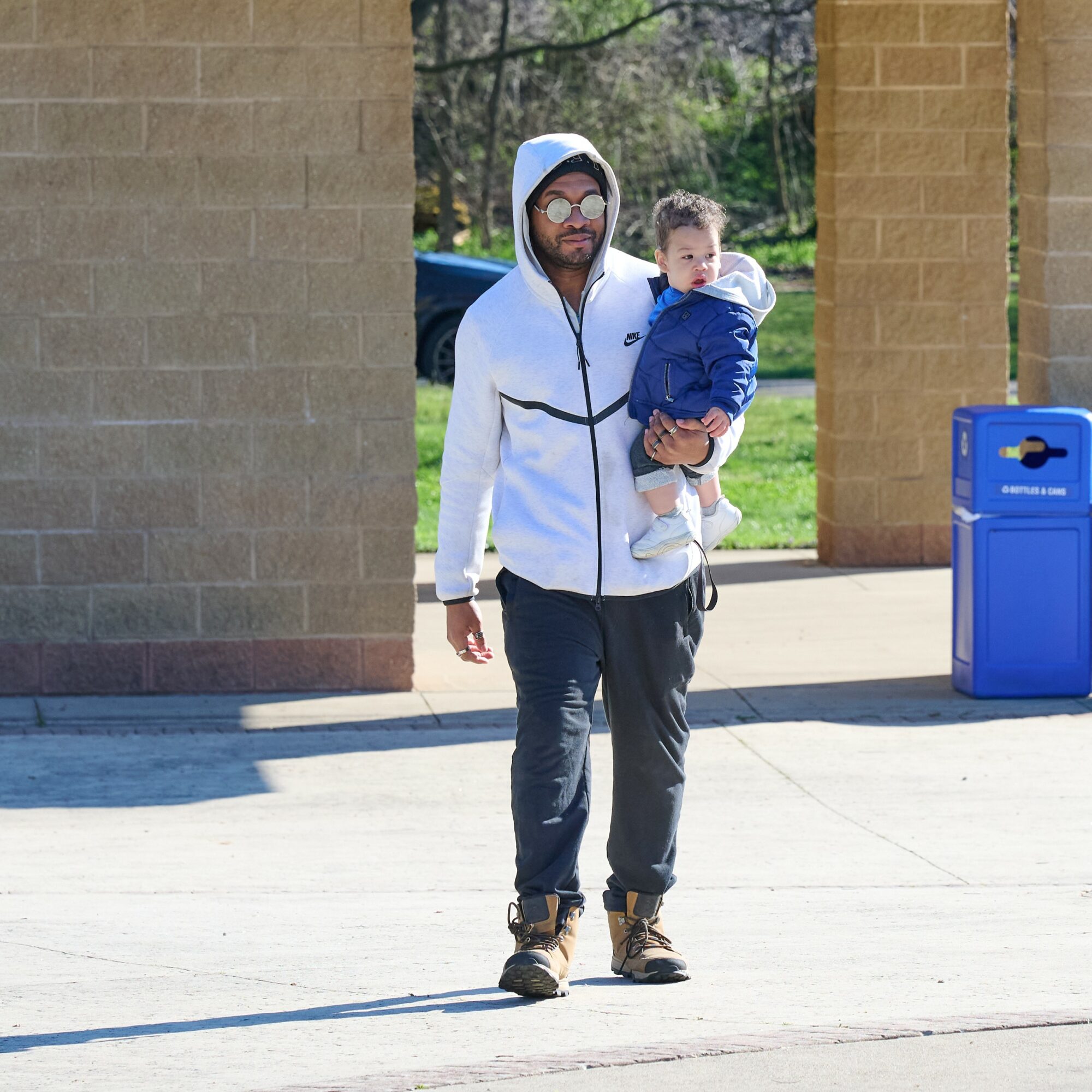 Man in white hoodie carrying a child in blue jacket outside, brick wall and blue trash bin behind them.