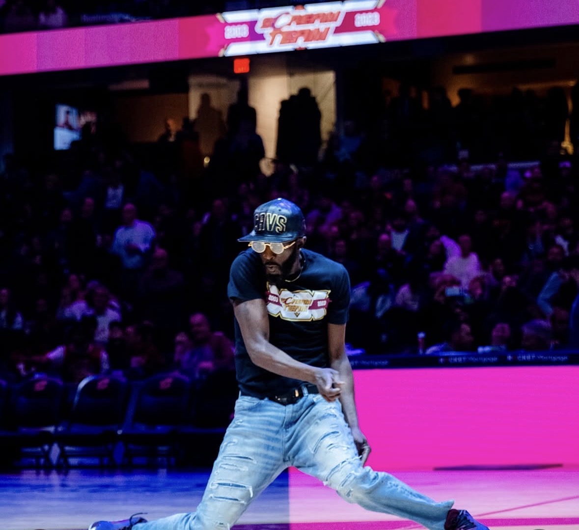 Person performing dance move on stage with audience in background, wearing cap, sunglasses, and casual clothing.