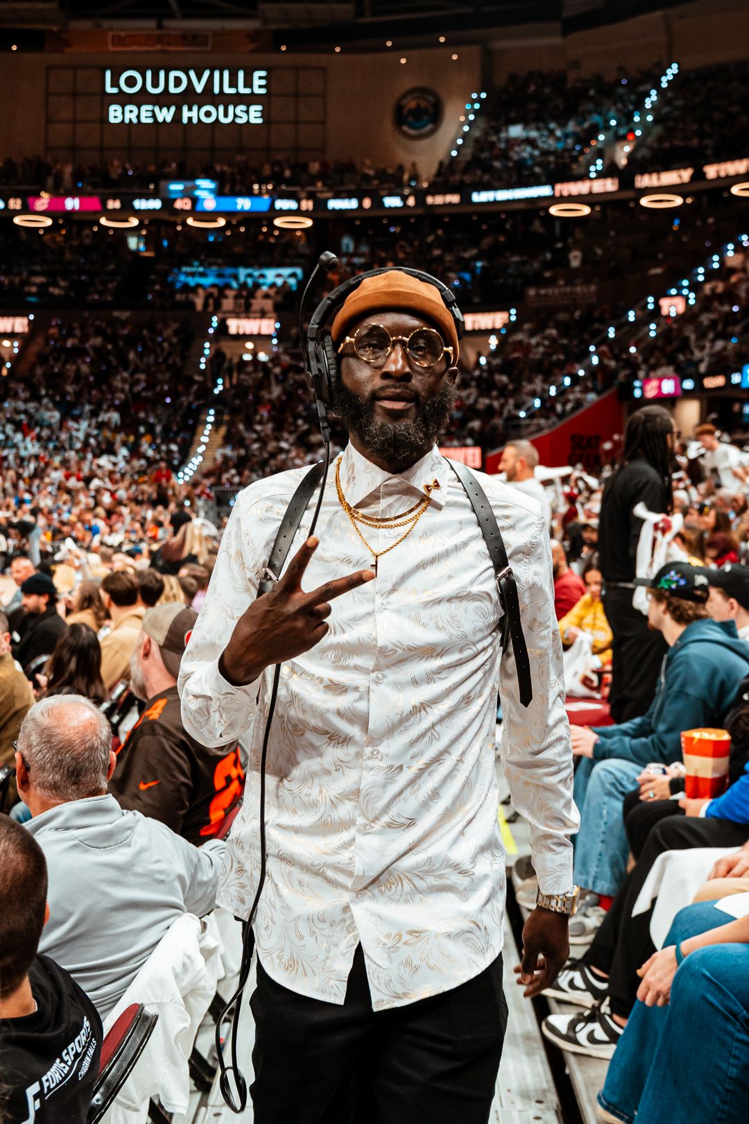 Man with glasses and a hat making a peace sign at a crowded indoor event, with a sign reading 'LOUVDVILLE BREW HOUSE' in the background.