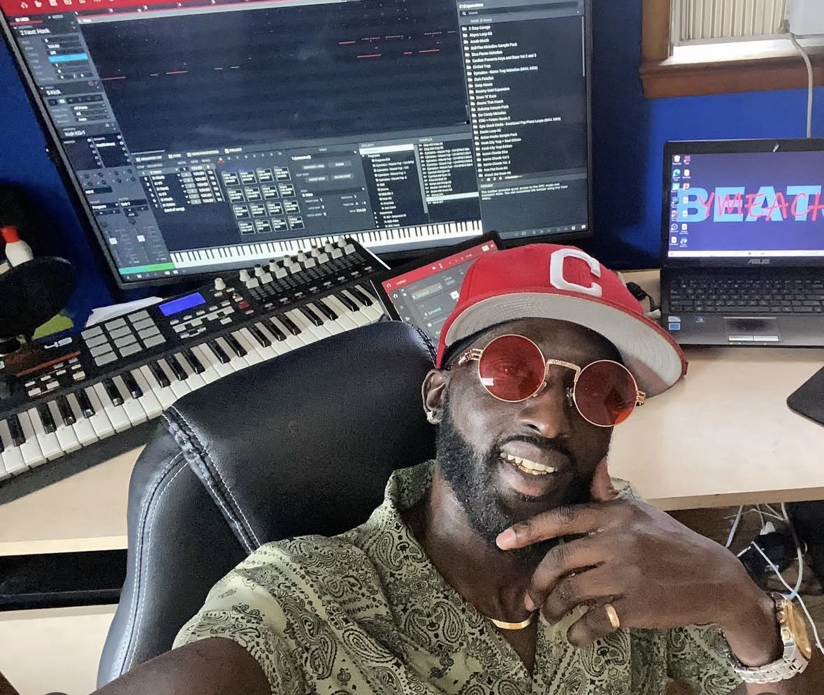 Man wearing sunglasses and a cap, sitting in a music studio with computers and musical equipment.