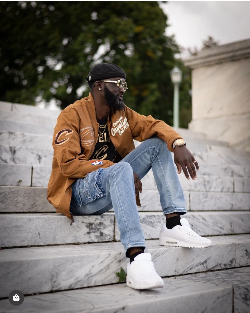 Man sitting on stone steps outdoors, wearing a brown jacket, jeans, white sneakers, sunglasses, and a black cap.