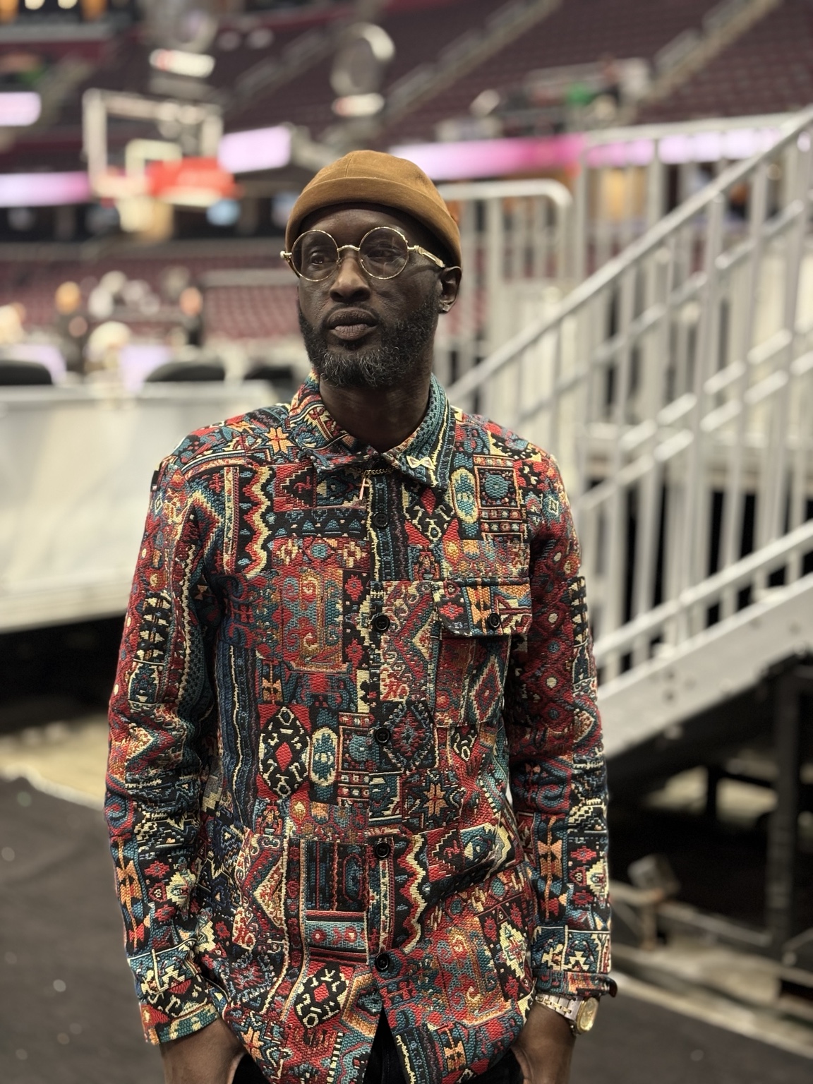 Man with glasses and a hat standing indoors, wearing a colorful patterned shirt, in front of stairs and seating area.