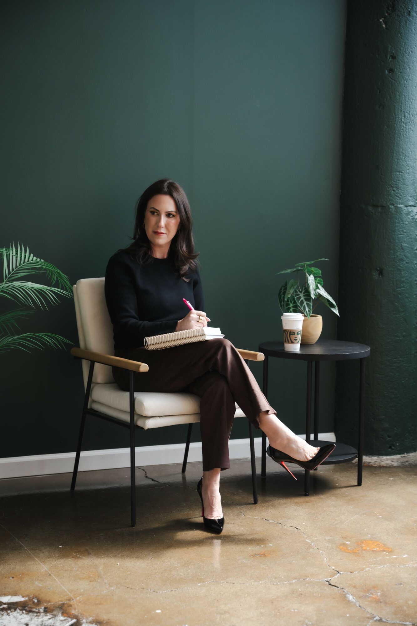 Woman sitting on a chair with a notebook, plants, and a coffee cup nearby, against a dark green wall.