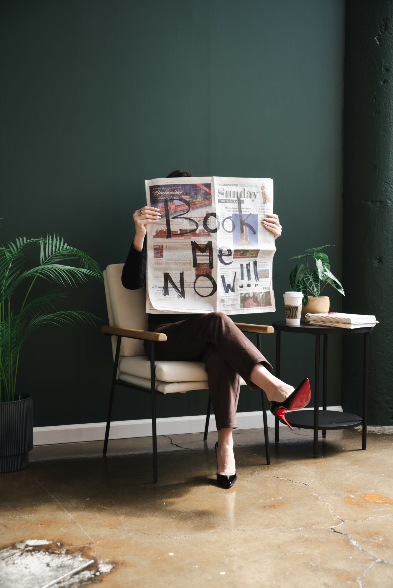 Person sitting on a chair reading a newspaper, legs crossed, in a room with green walls and plants.