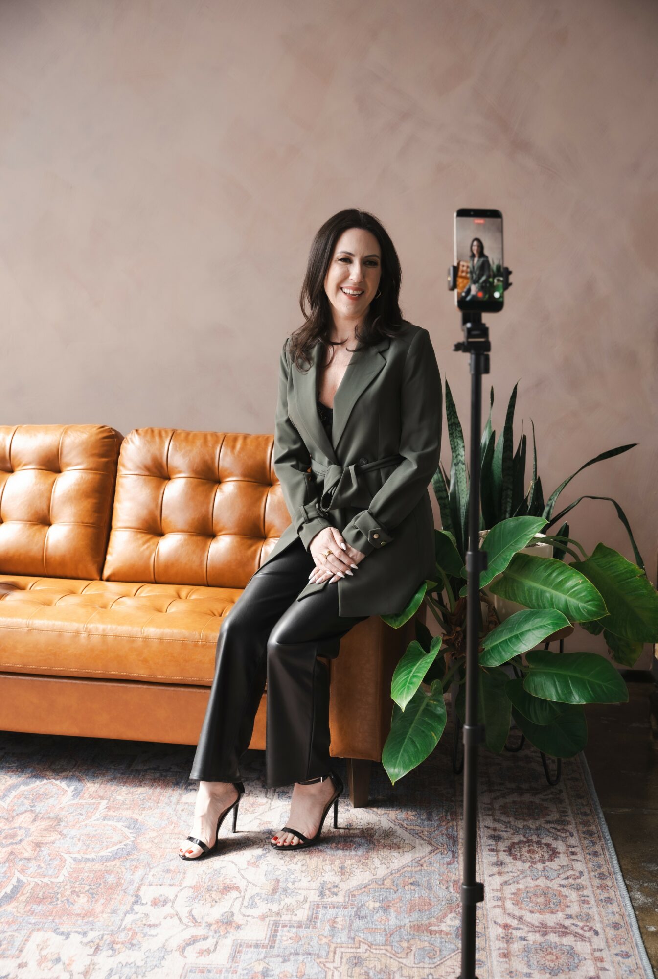Woman sitting on a tan leather sofa, smiling, with a smartphone on a tripod, next to large green plants, in a room with a beige wall.